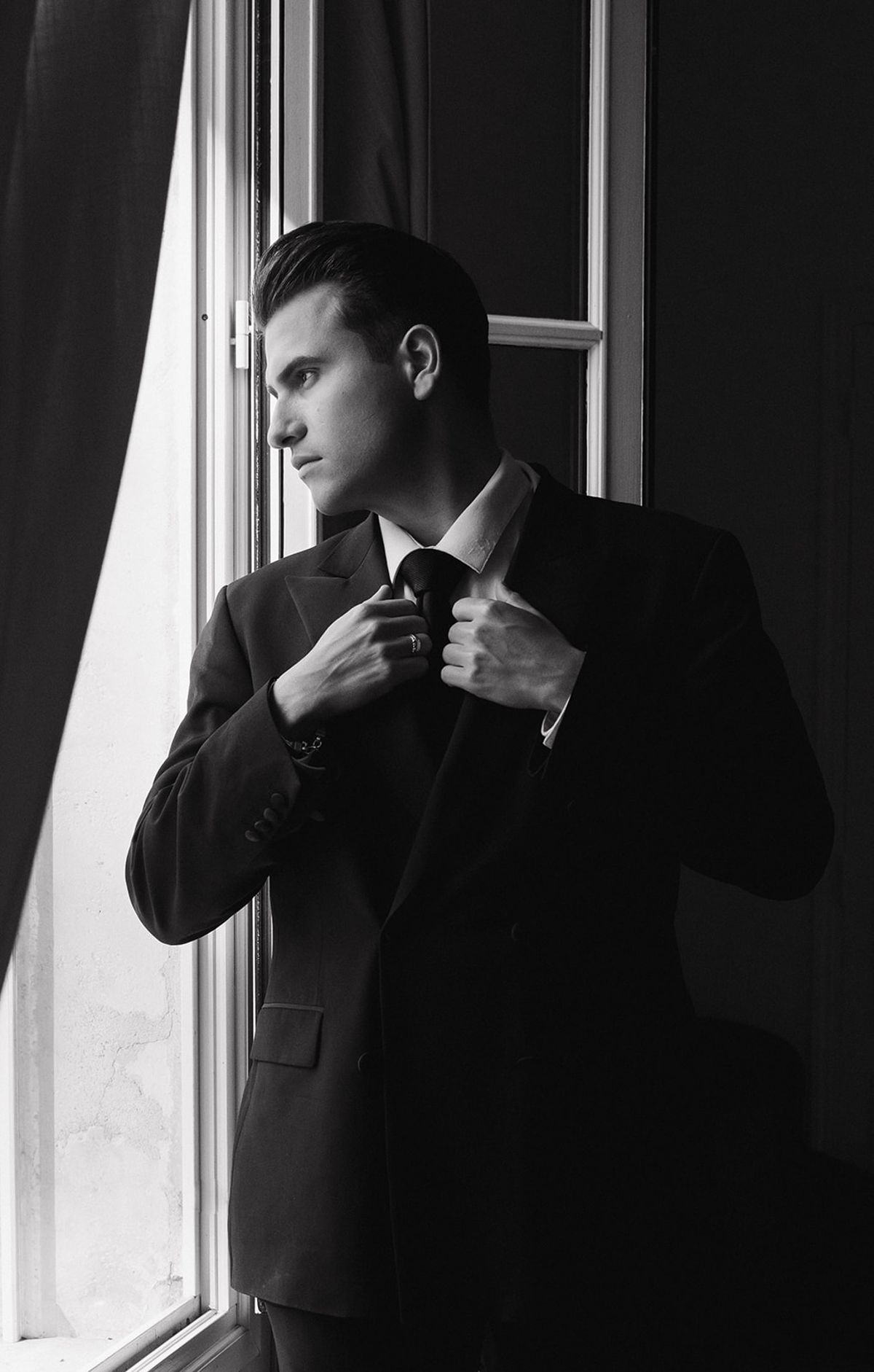 Black and white portrait of the groom adjusting his jacket at a window