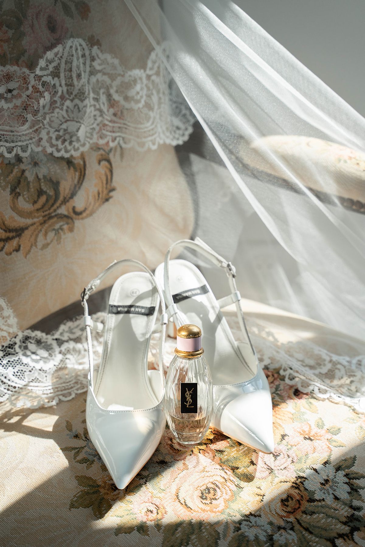 Bridal shoes and a perfume bottle arranged on embroidered fabric beside the veil