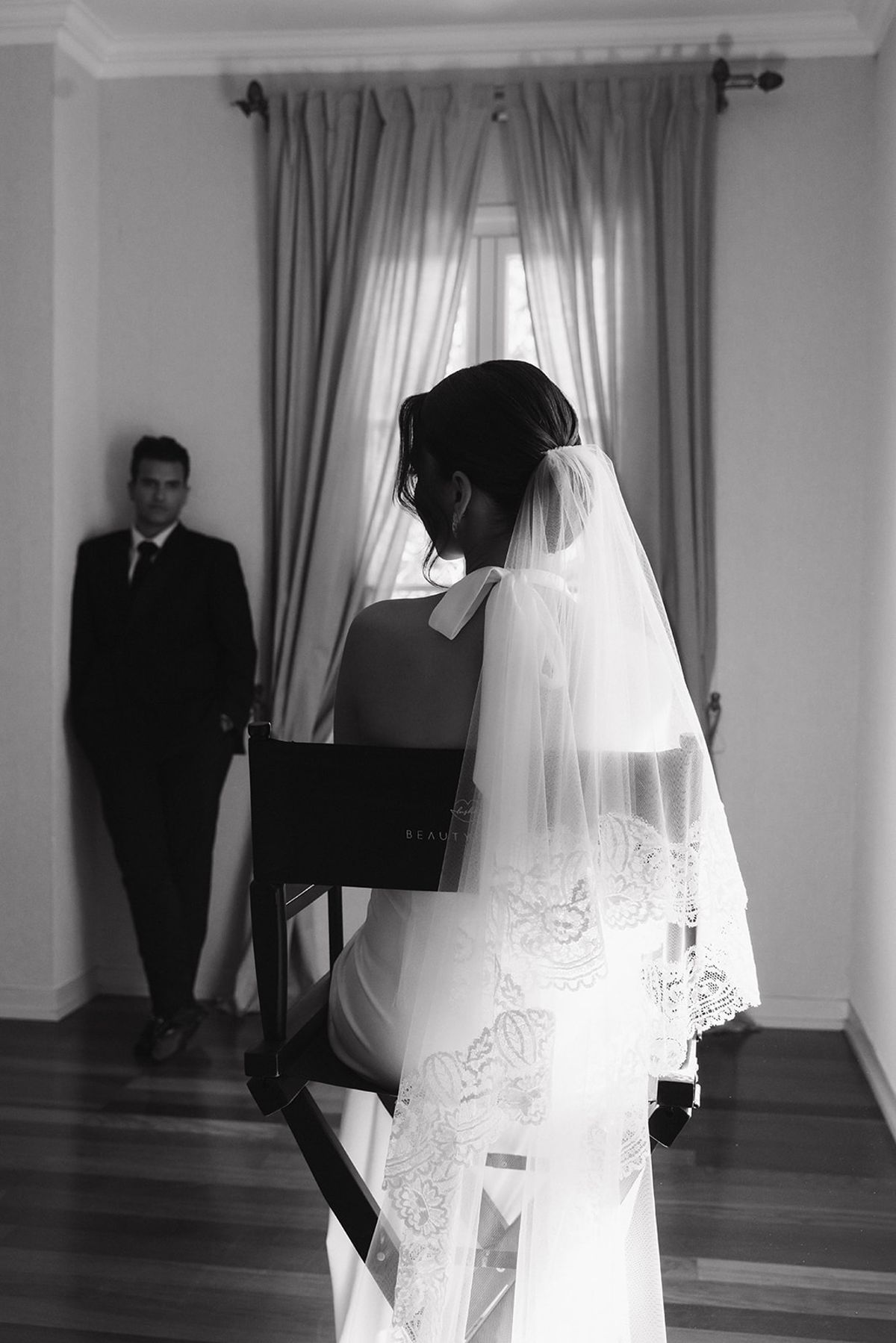 Black and white image of the bride seated in a makeup chair with the groom waiting behind