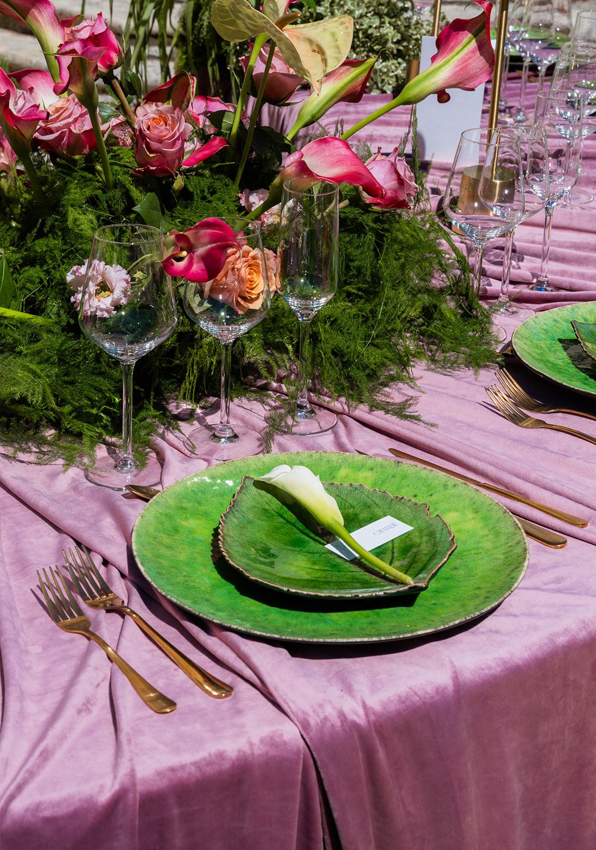 Place setting with a deep green glass plate, gold cutlery and a single calla lily on mauve velvet