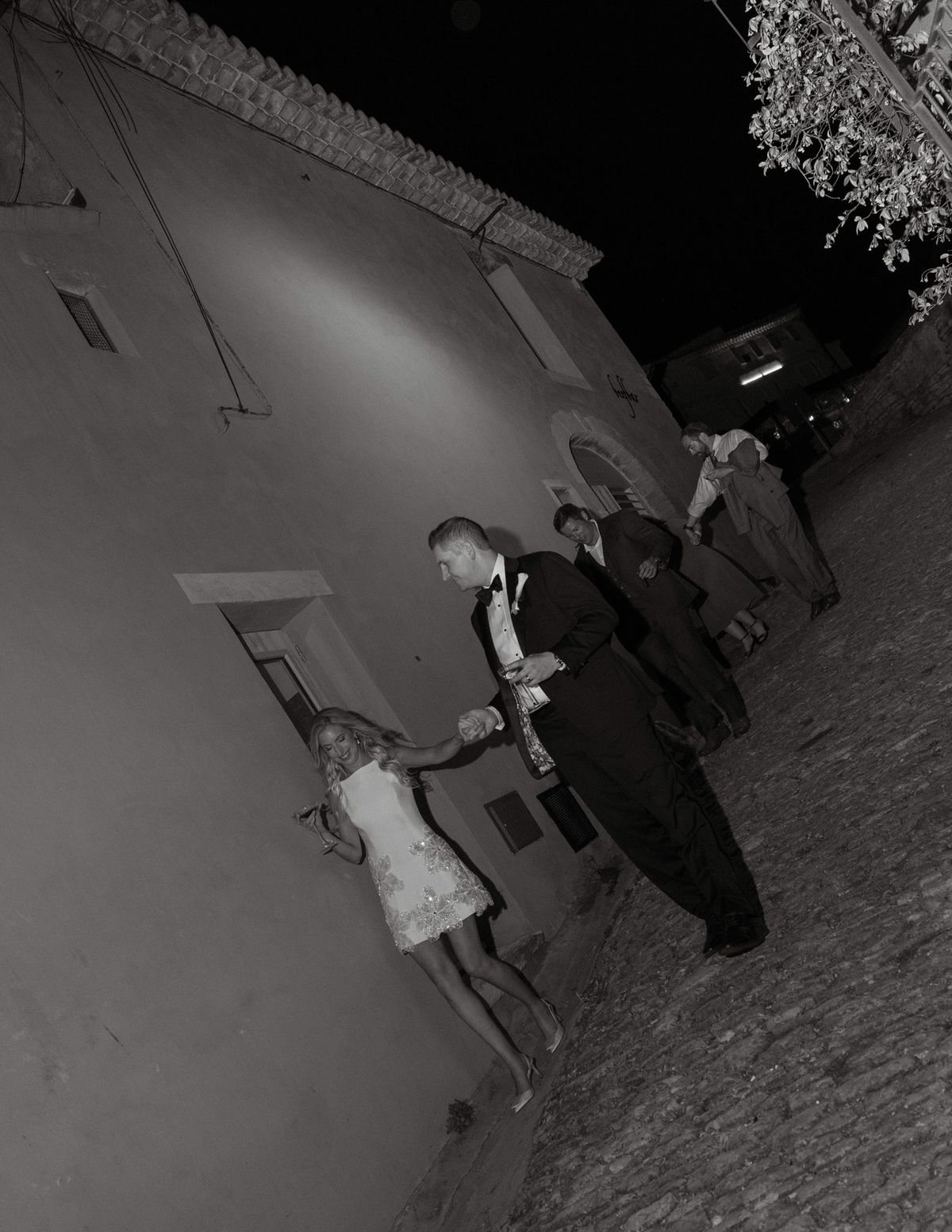 Black and white photo of Ryan leading Kirstin in her short dress along a village street at night