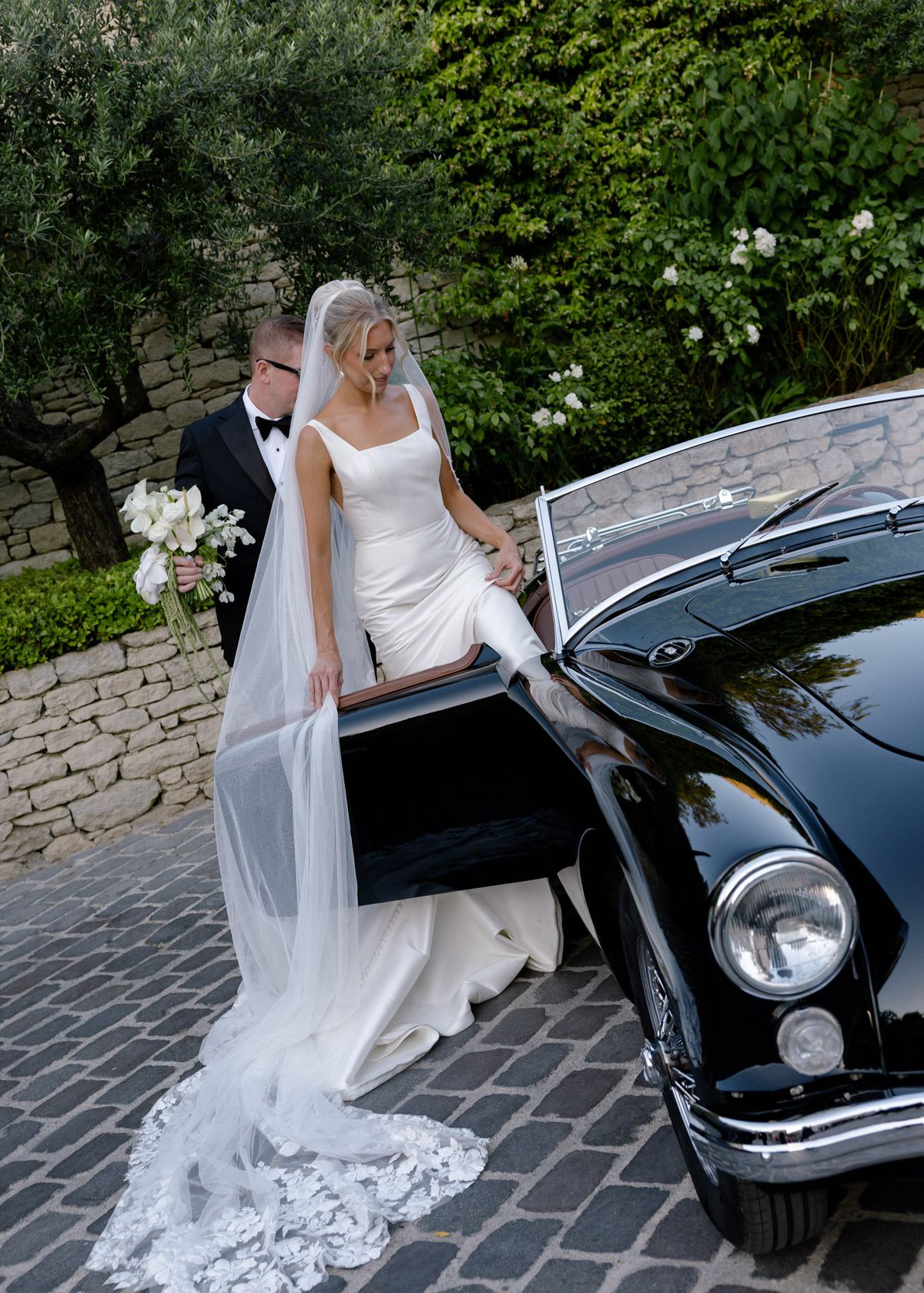 Bride stepping into a black vintage convertible as the groom holds her bouquet