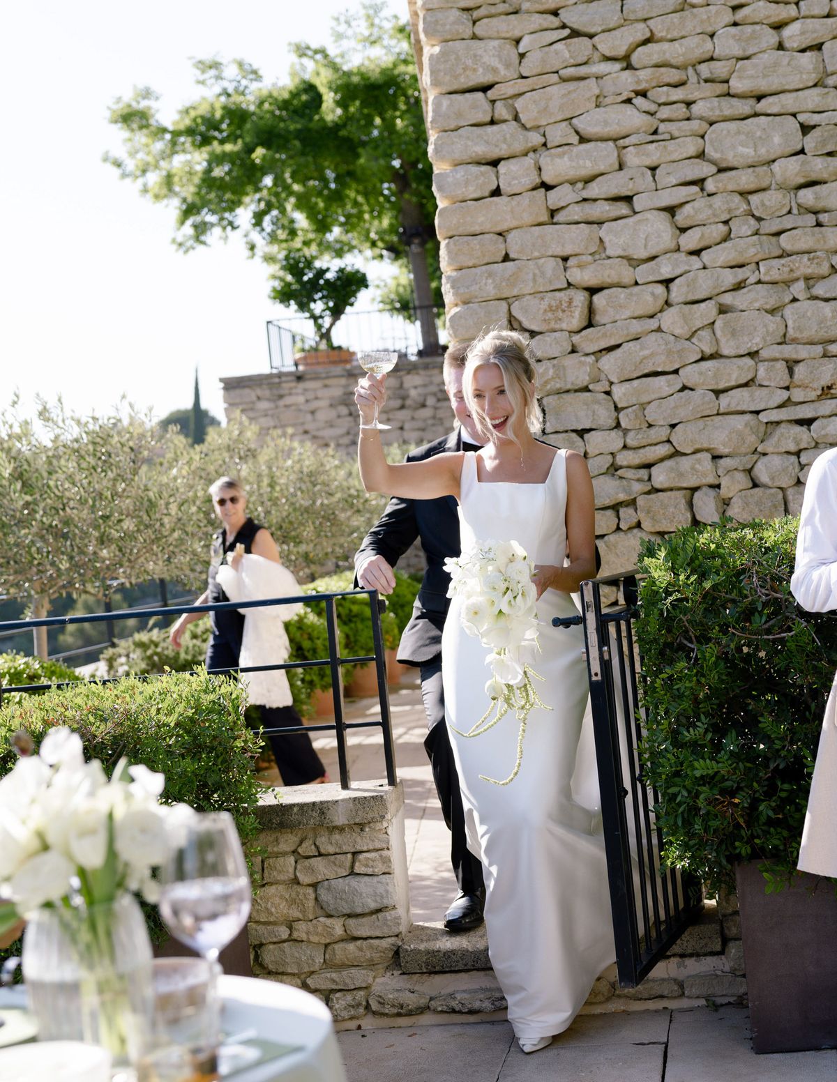 Kirstin raising a champagne coupe as she and Ryan come through a gate onto the terrace