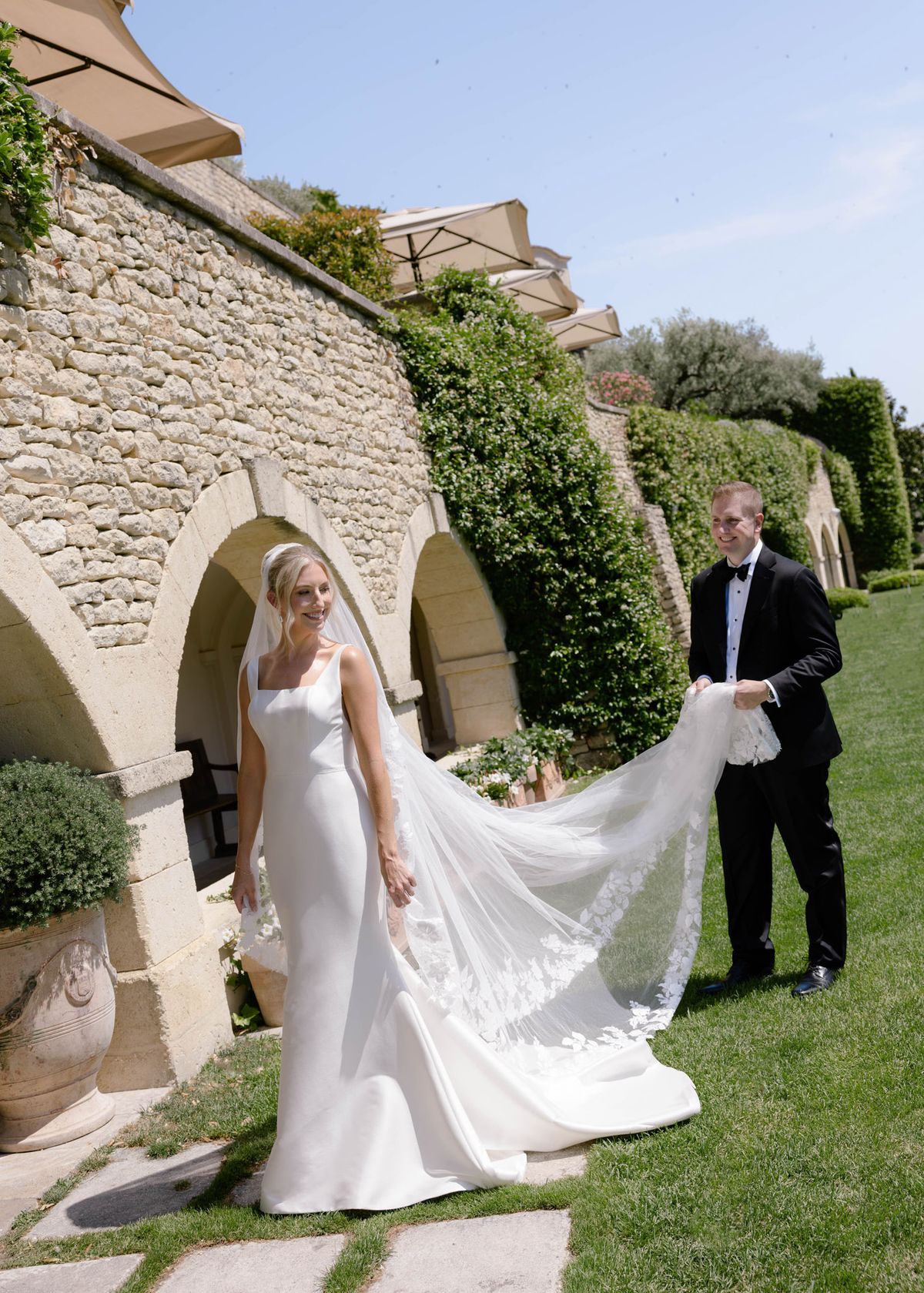 Groom holding the bride's train beneath a stone arcade