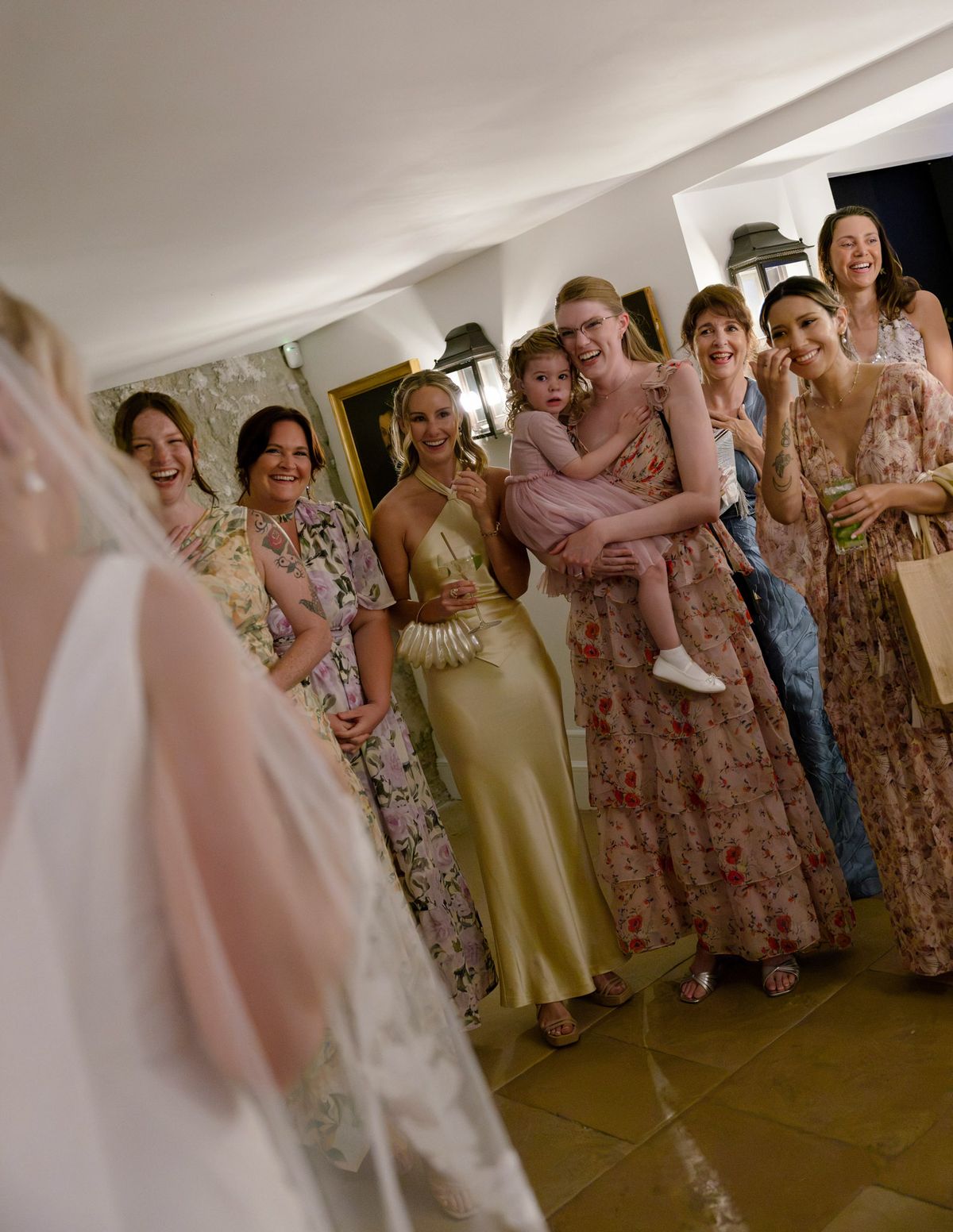 Friends in floral dresses reacting as they see the bride, her veil in the foreground