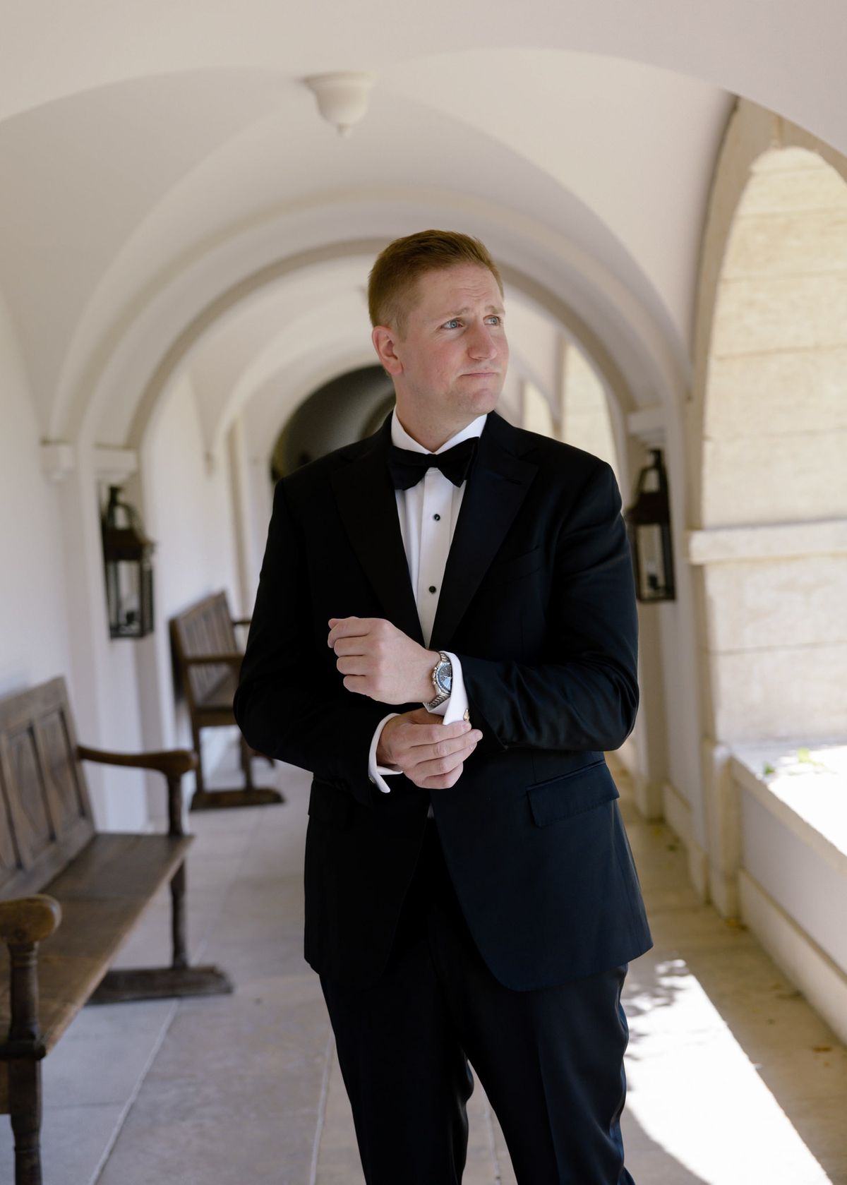 Groom in a tuxedo fastening his cuff in a stone arcade