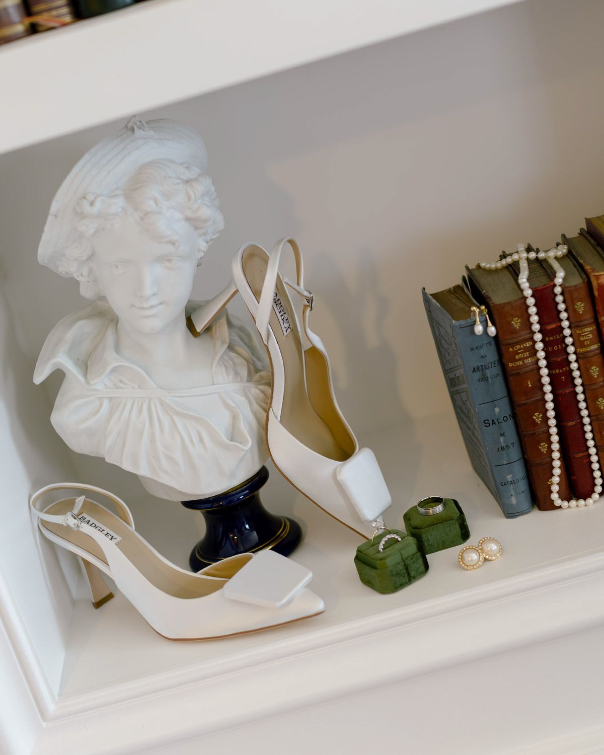 Bridal heels, pearls and wedding rings on a shelf beside a white bust