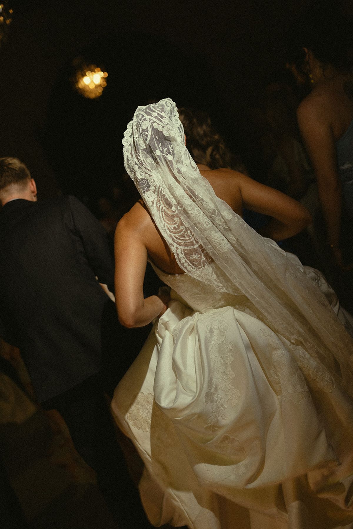 The bride, seen from behind in a long lace veil and full-skirted gown, moves through a dimly lit crowd of guests.