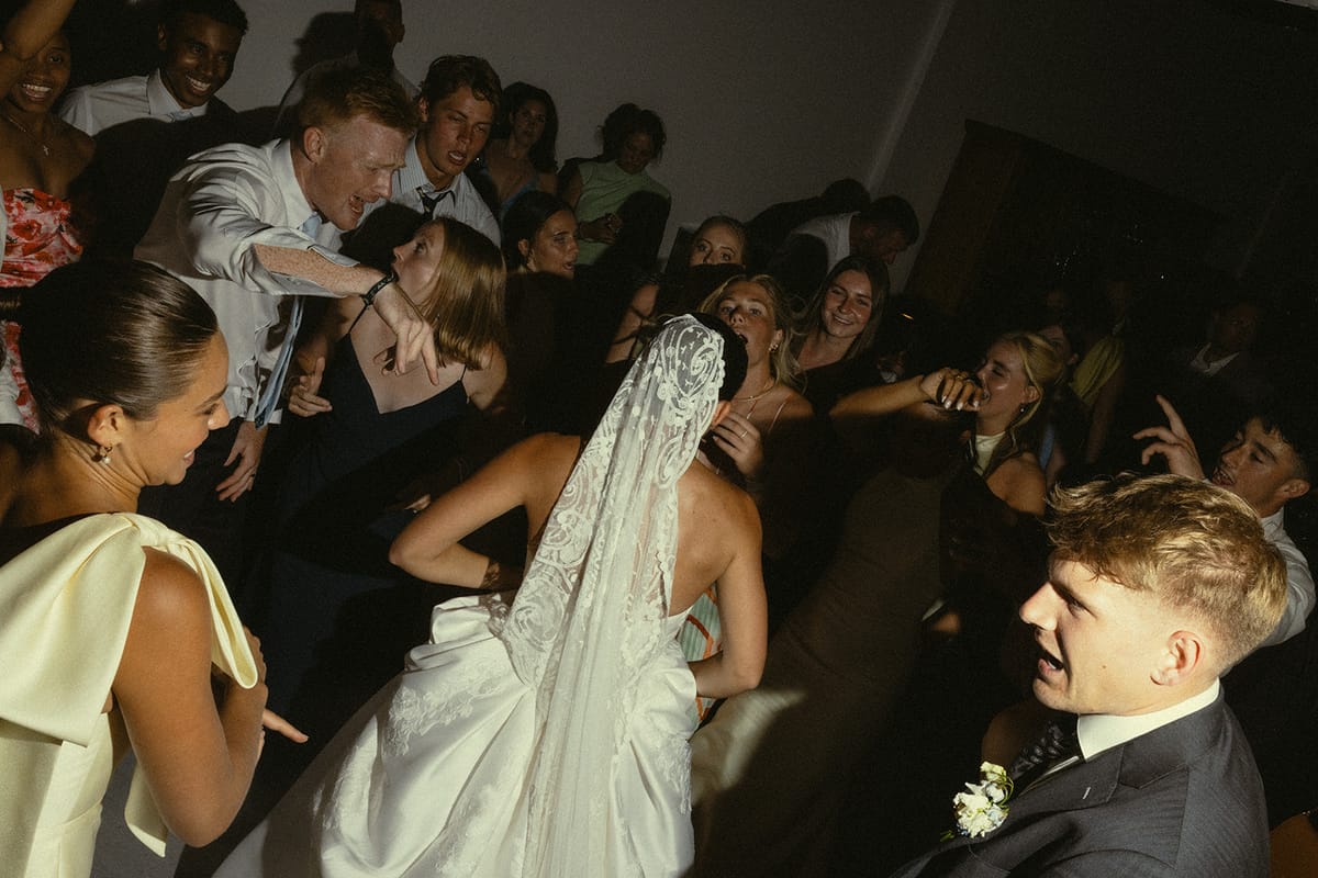The bride in a veil and the groom are surrounded by a crowd of guests in a dimly lit room.