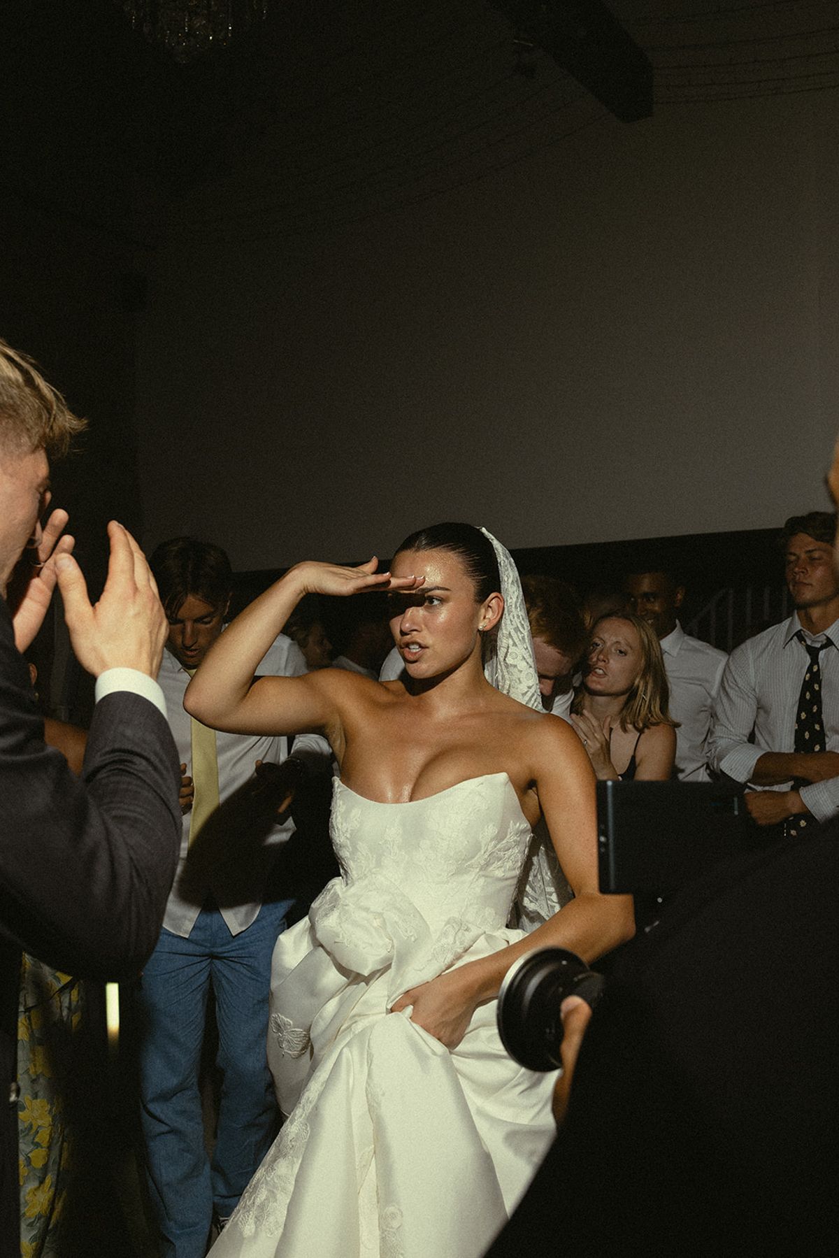 The bride in a white gown raises a hand to shade her eyes while guests and a photographer surround her in a dark room.