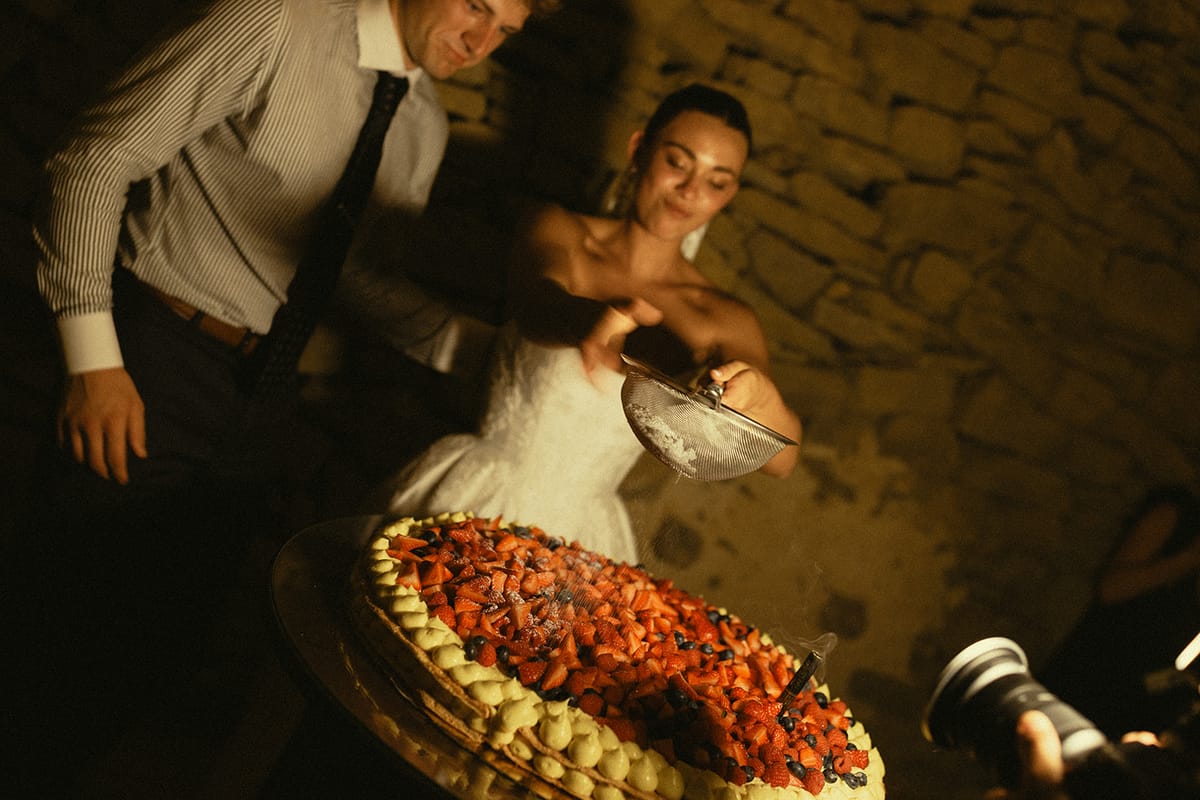 The smiling bride holds a small sieve over a dessert topped with red berries while a man stands beside her in low light.