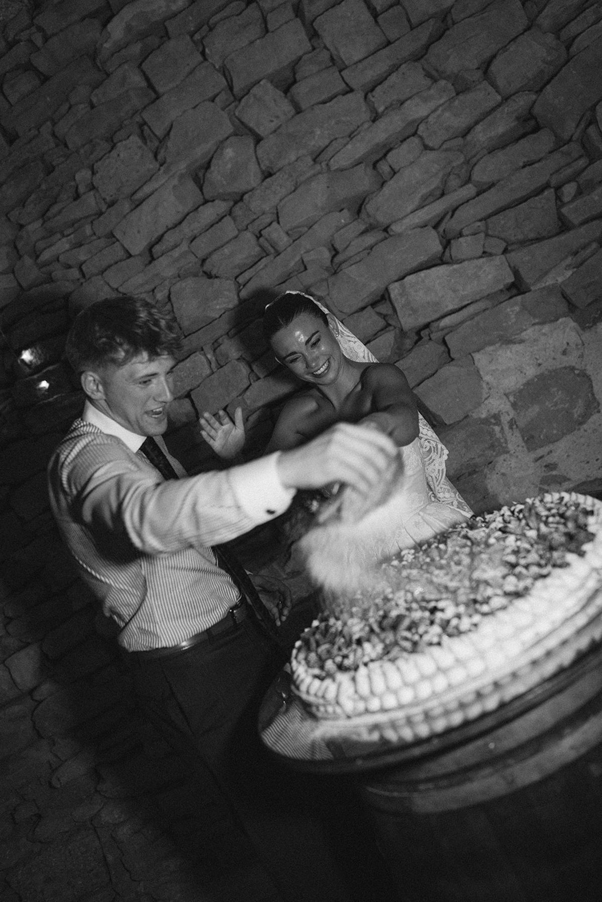 In black and white, the couple lean over a large cake to cut it, in front of a rough stone wall.