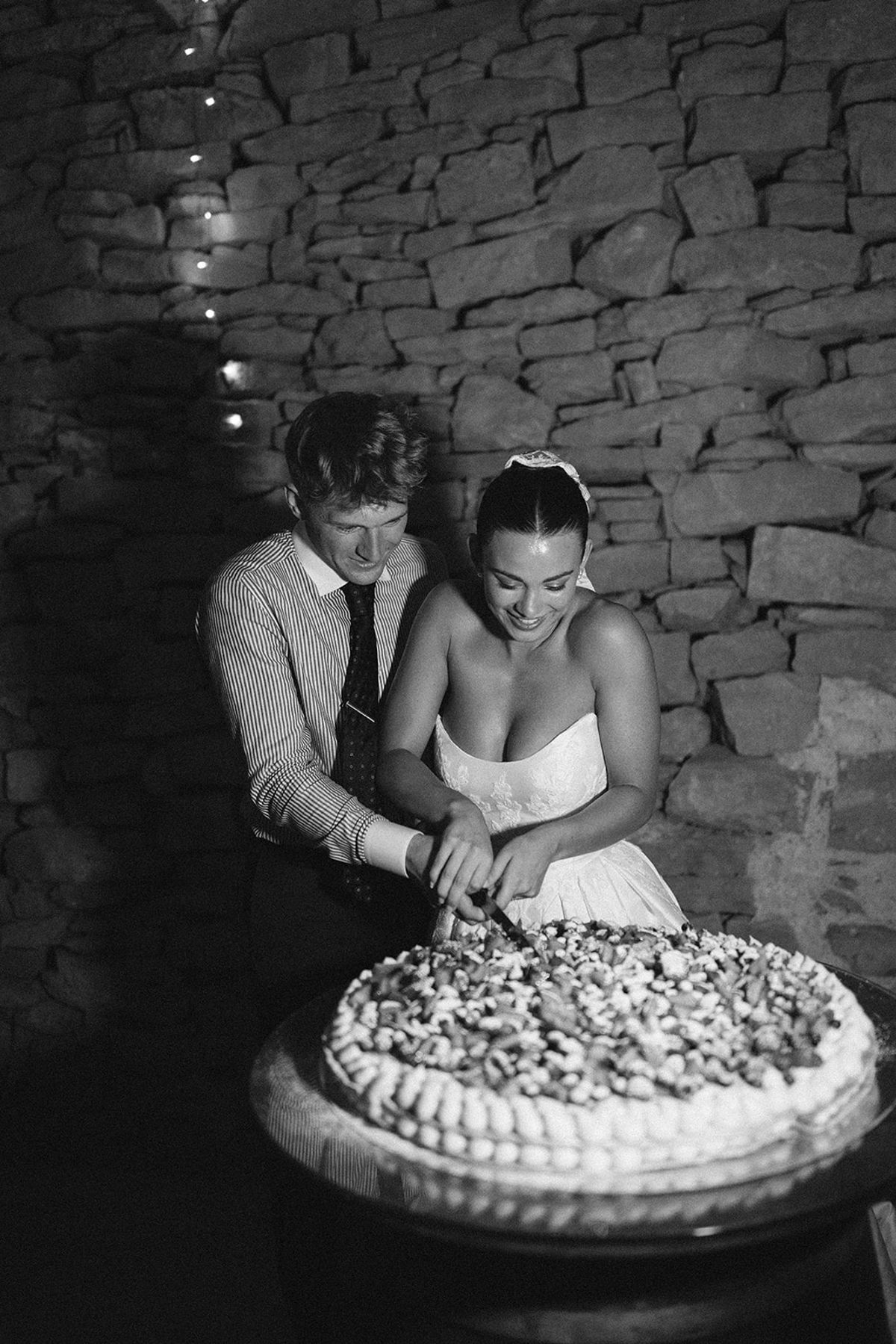 In black and white, the couple cut a large round tart together against a rough stone wall.