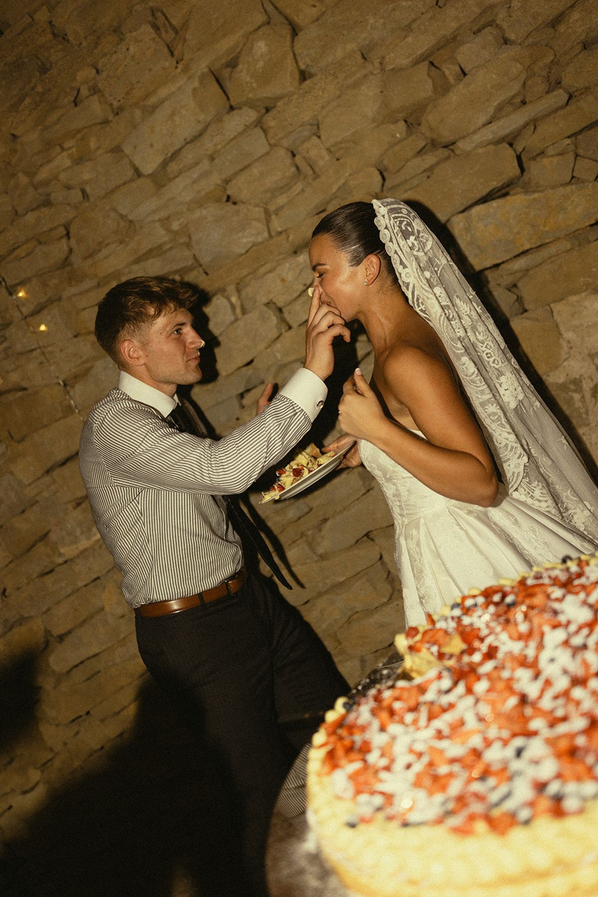 The groom in shirtsleeves feeds cake to the bride in her veil, with a decorated cake in the foreground.