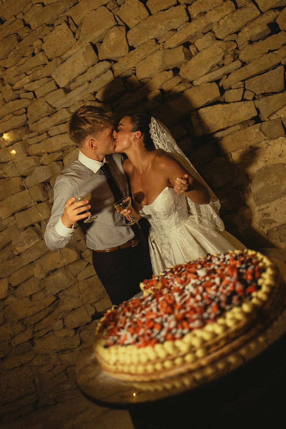 The couple kiss against a rough stone wall, holding glasses, with a tart topped with red berries in the foreground.