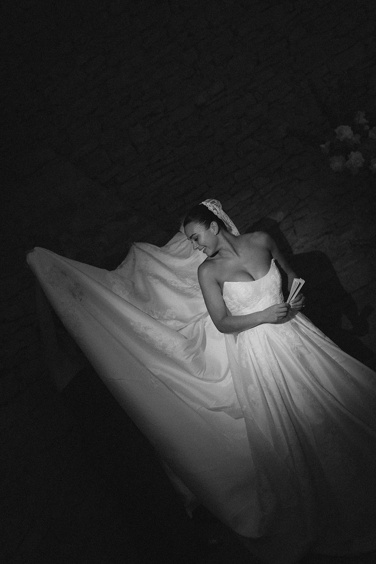In black and white, a bride sits with her head bowed as her voluminous white gown spreads around her in the darkness.