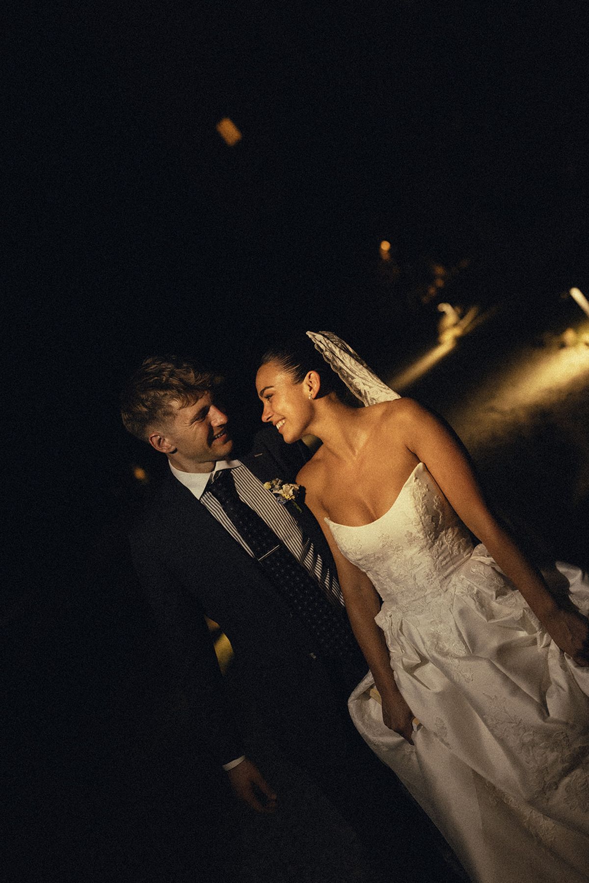 In low light, the groom in a dark suit and the bride in a strapless gown and veil smile at each other.