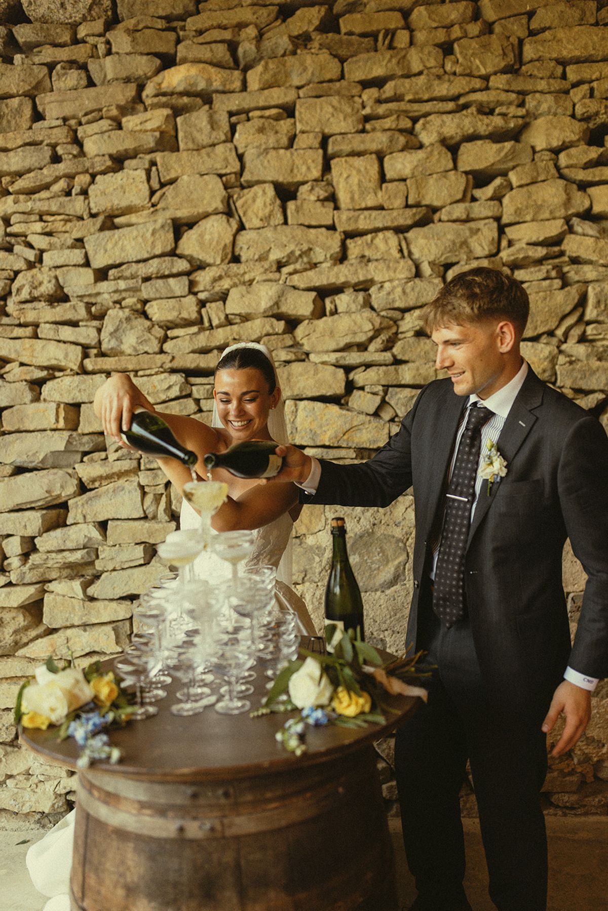 The bride and groom pour champagne into a pyramid of glasses set on a wooden barrel in front of a rough stone wall, with flowers beside it.