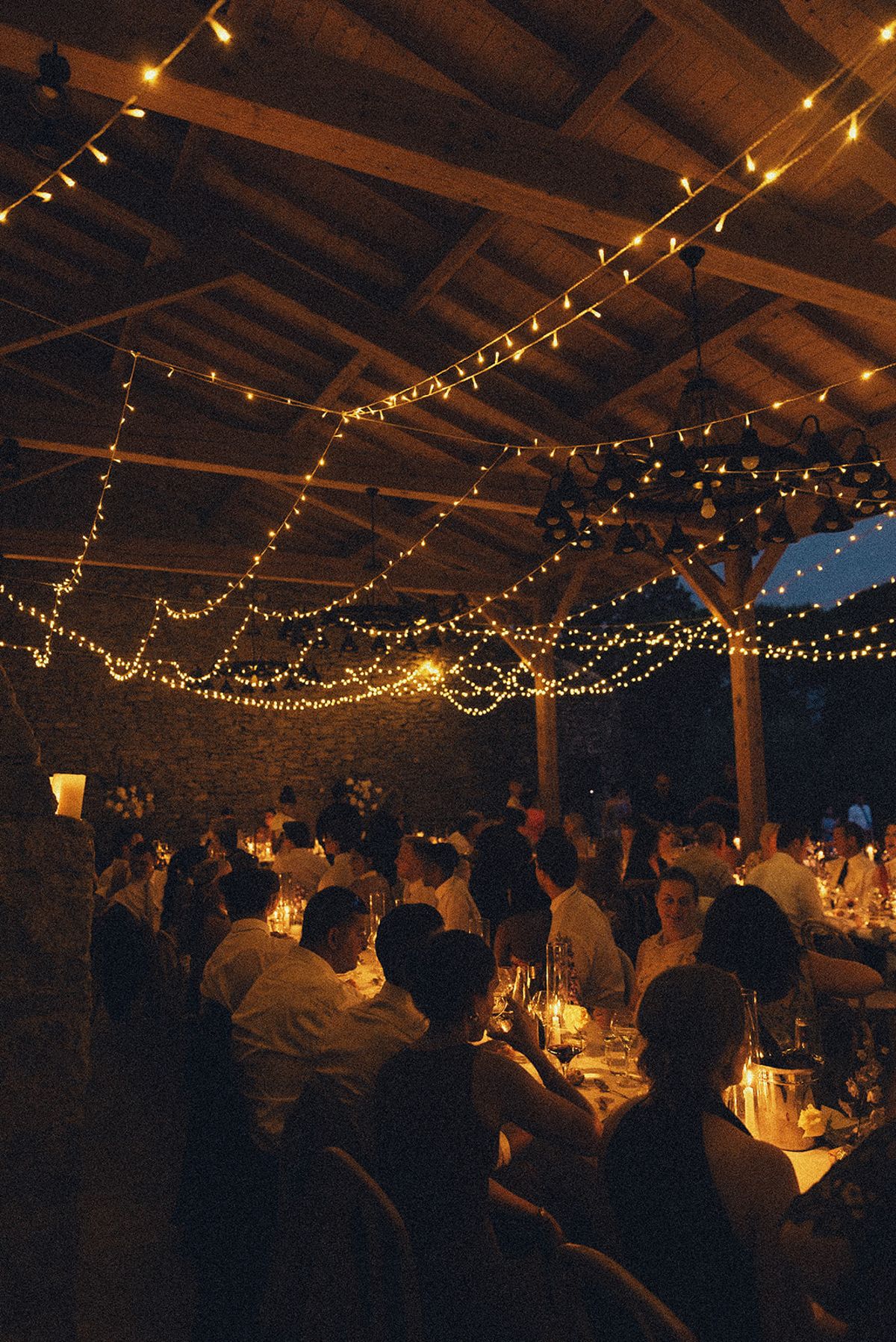Guests sit at long candlelit tables in a timber-beamed hall hung with strings of warm lights.