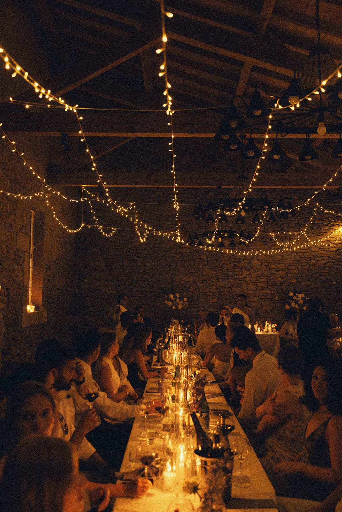Guests sit along a long table lit by candles, beneath wooden beams hung with strings of small lights.
