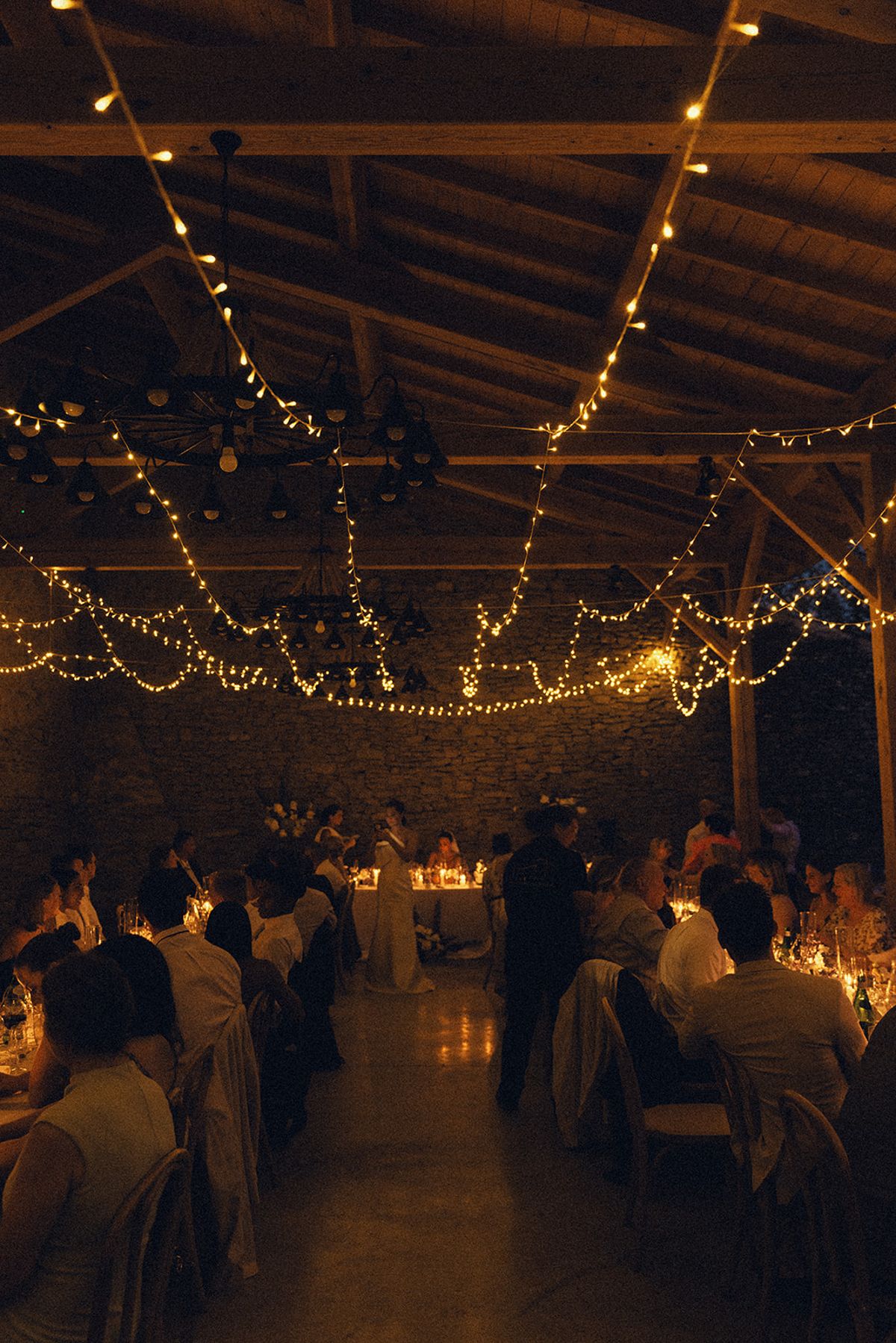 Guests sit at tables in a wide timber-beamed hall, lit by candles and overhead strings of lights.