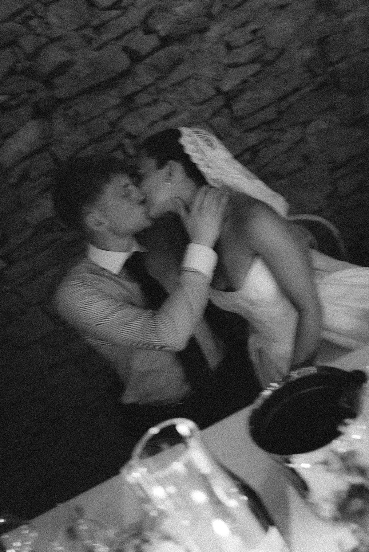 In black and white, the couple kiss closely, the bride's veil beside them and drinking glasses in the foreground.