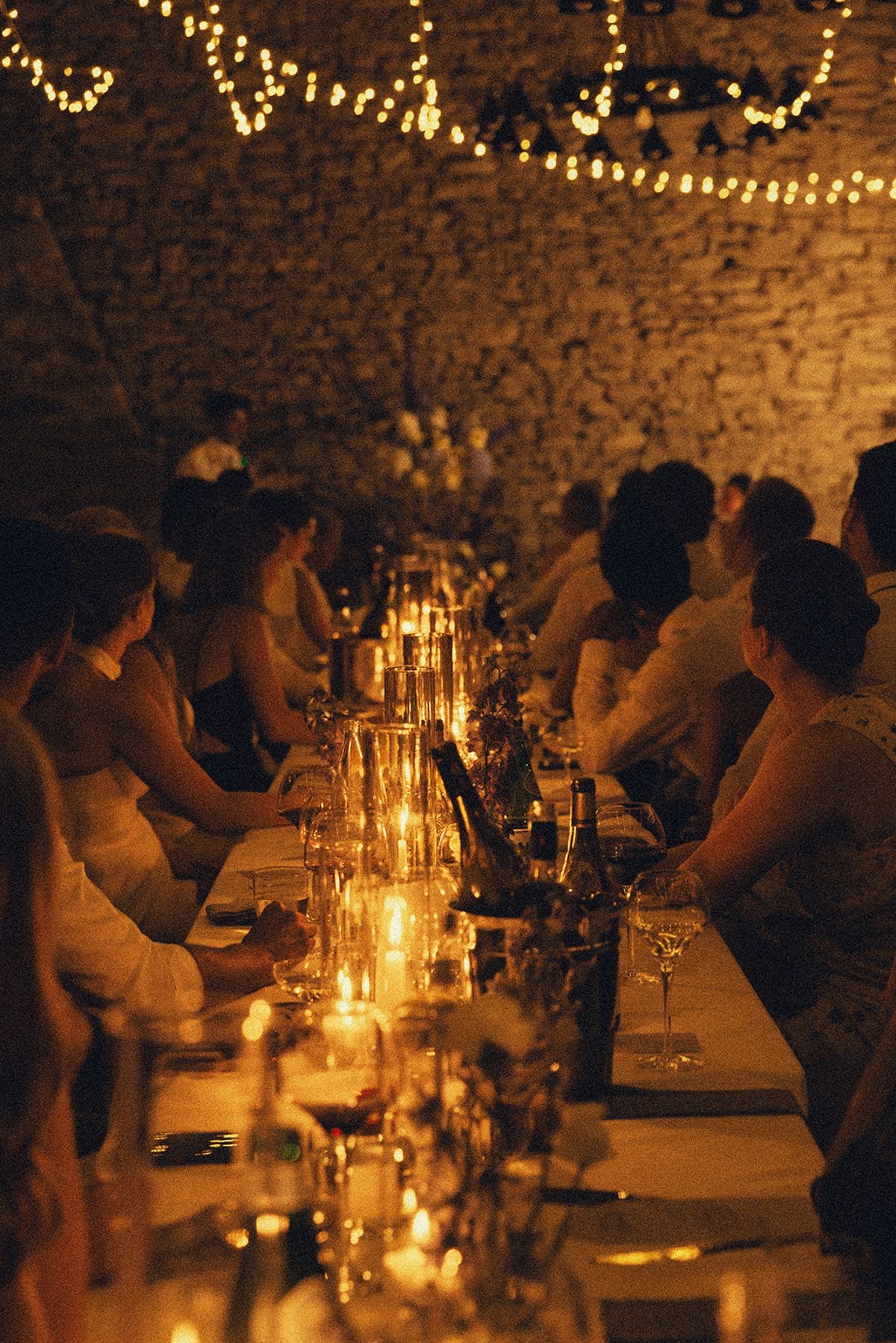 Guests sit along both sides of a long table lit by many candles, beneath strings of fairy lights.