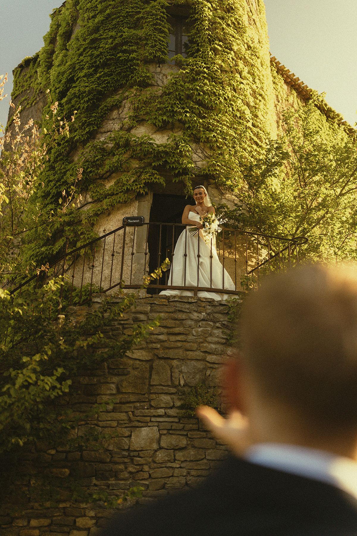 The back of the groom's head is in the foreground while the bride stands on stone steps by an ivy-covered tower in warm light.