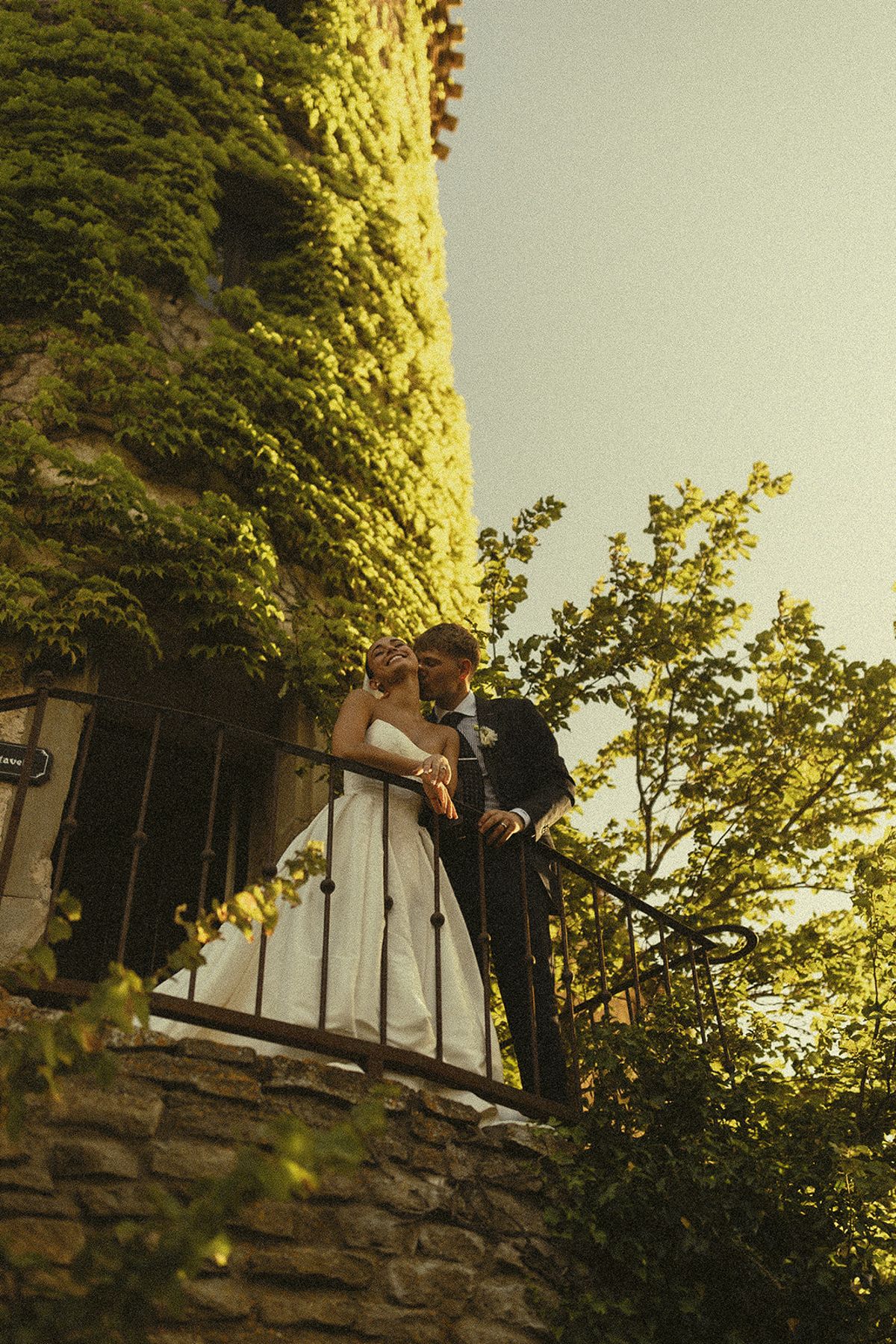 Seen from below, the couple embrace on a stone balcony of a tower covered in ivy, under a clear sky.