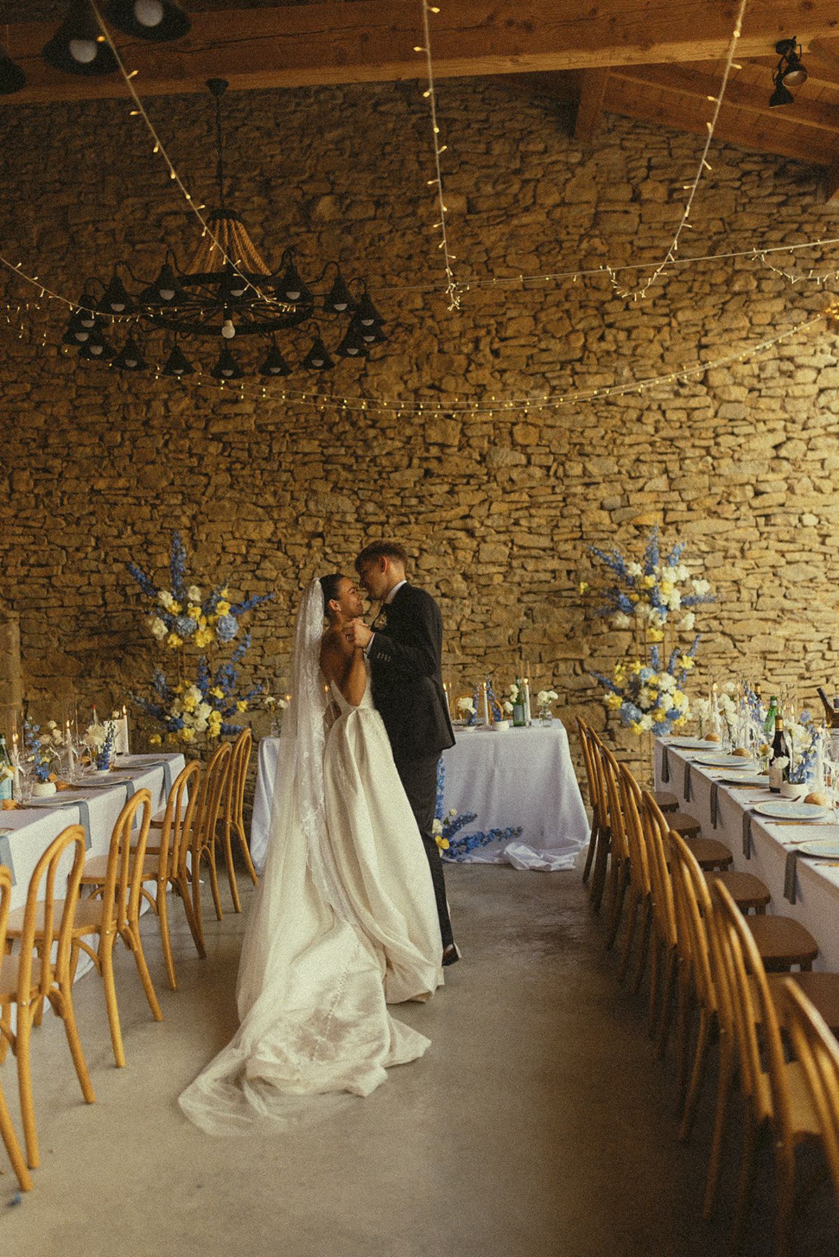 The couple embrace in the middle of a stone barn, between long set tables with blue and yellow flowers and wooden chairs.