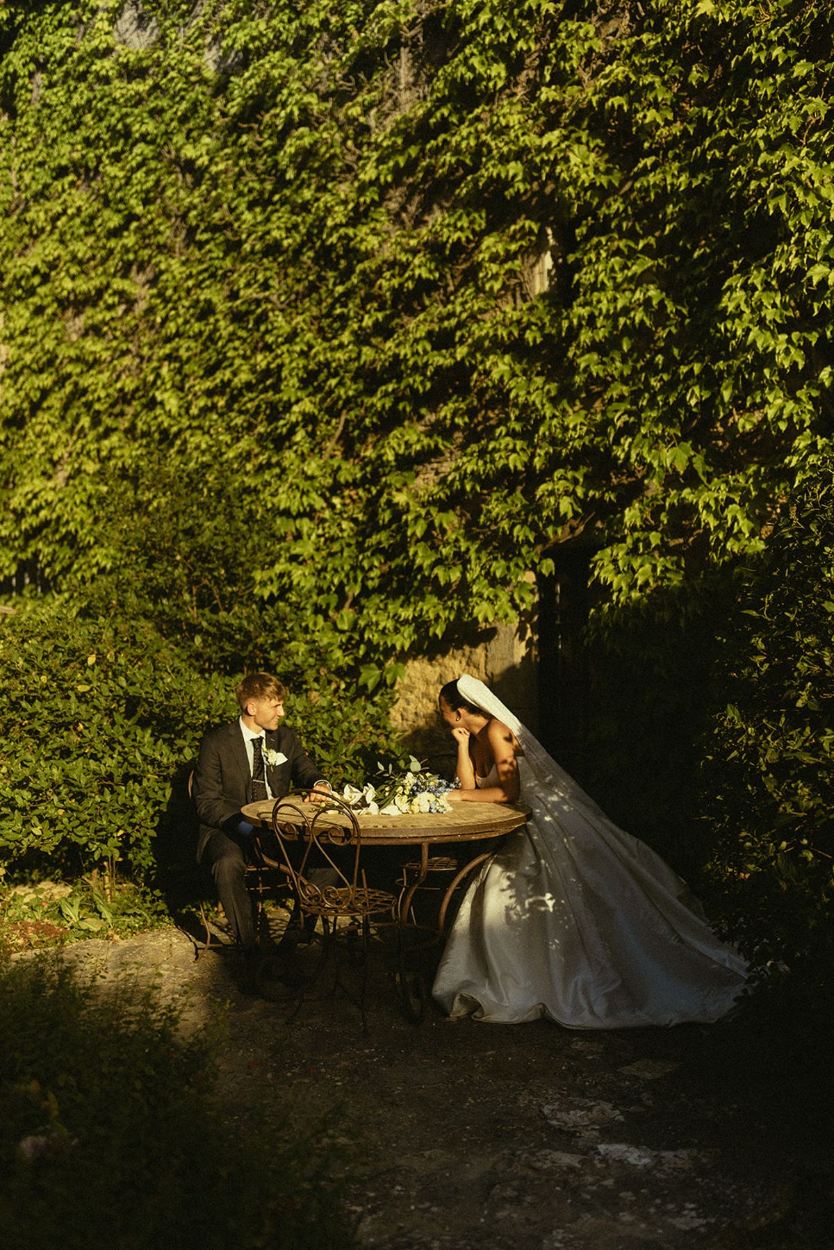 The couple sit at a small round table in a shaded garden surrounded by dense green ivy.