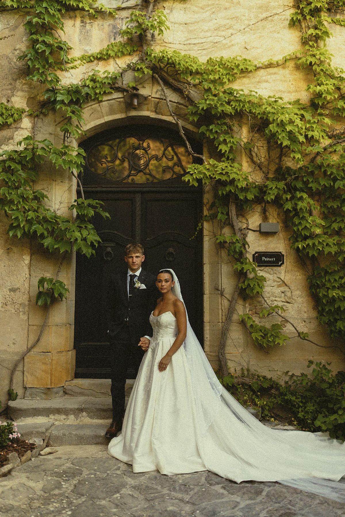 The groom in a dark suit and the bride in a white gown with a long train stand before a dark door framed by ivy.