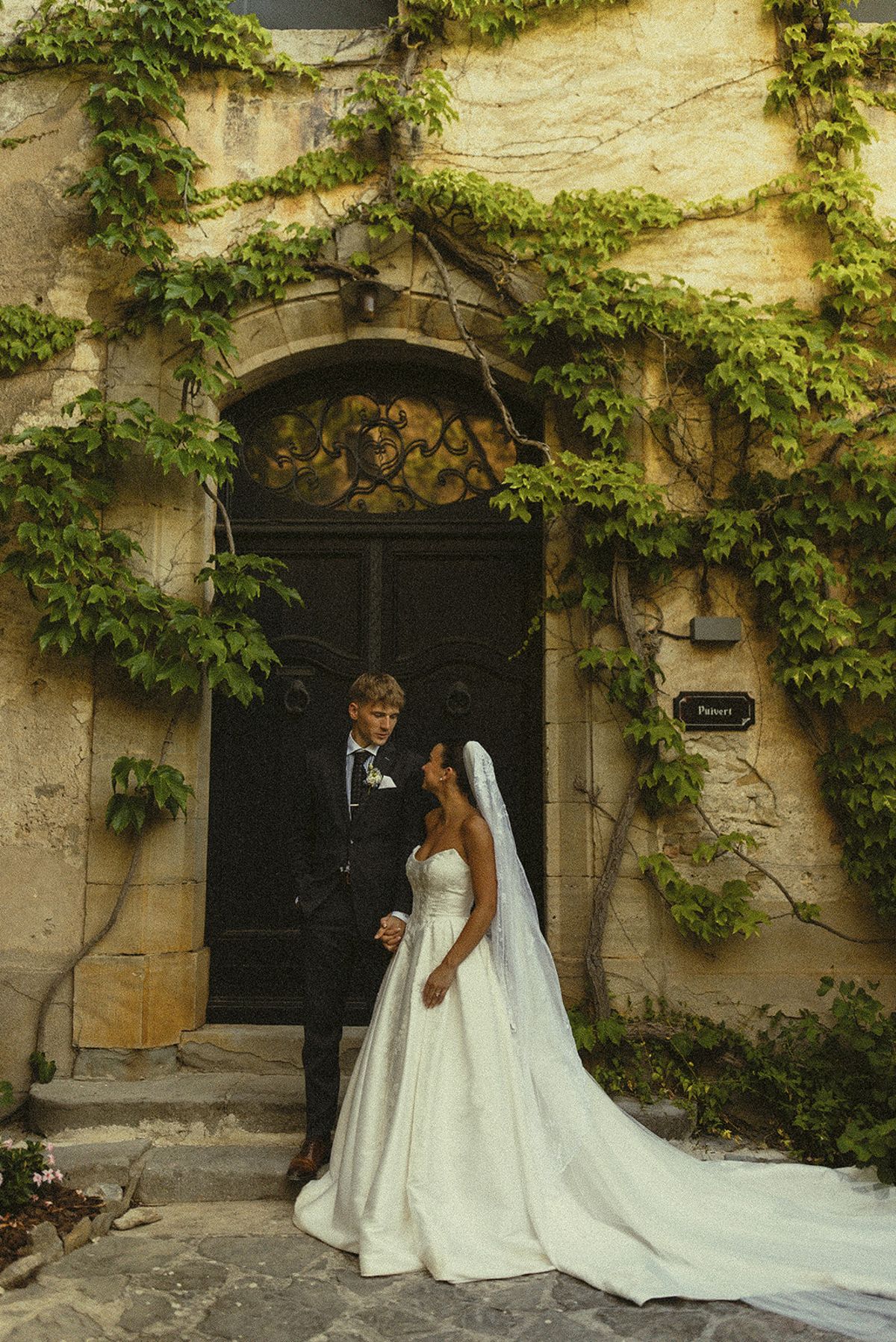 The couple stand beside a dark wooden door framed by green ivy on a stone wall, the bride's long veil and train trailing across the ground.