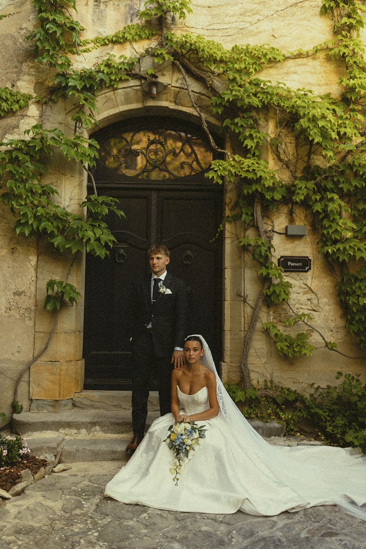 The groom stands in a dark suit while the bride sits on the steps beside him, her gown spread out before an ivy-framed door.