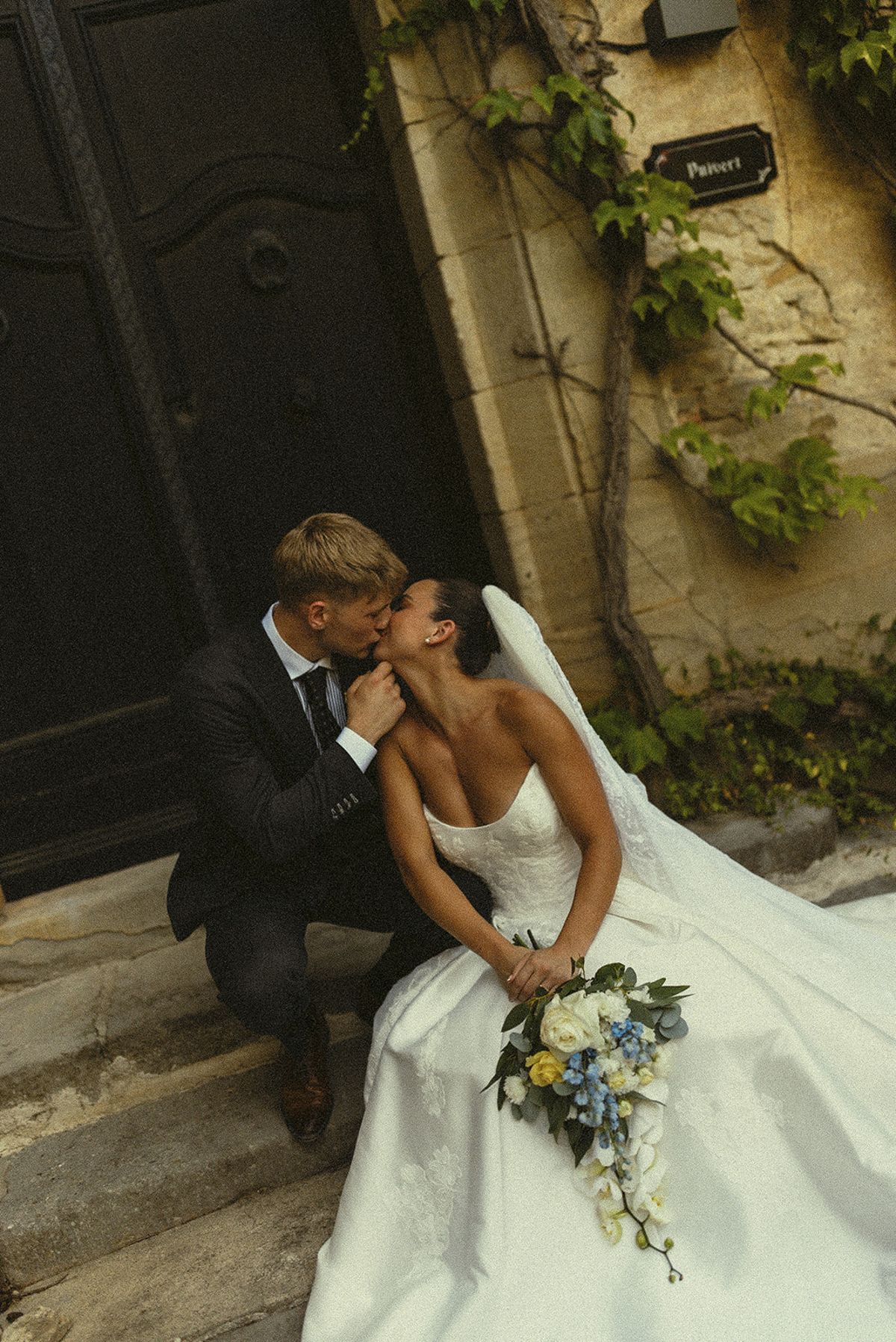 The couple kiss on worn stone steps beside a dark wooden door, the bride holding a bouquet of white and blue flowers.