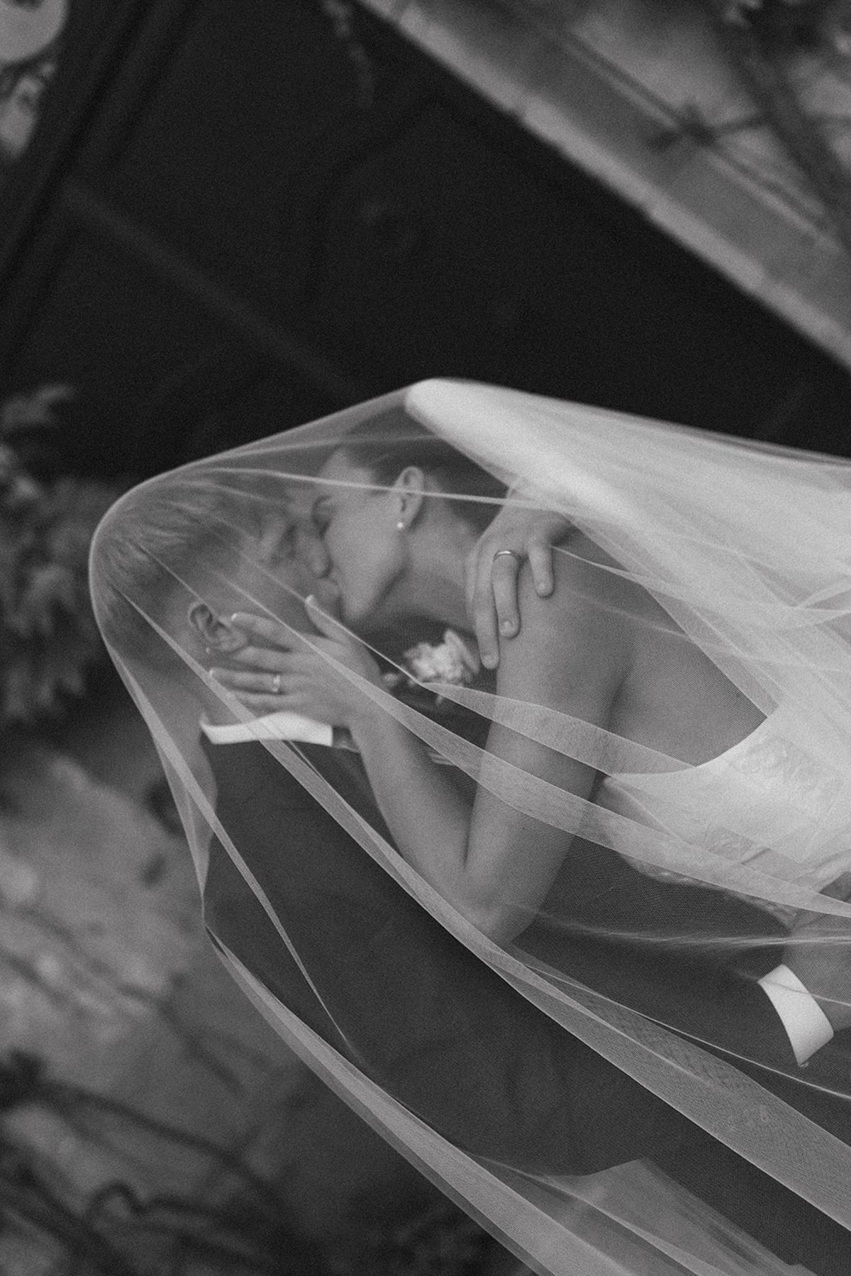 In black and white, two people embrace closely beneath a sheer veil that drapes across the frame, their faces softly visible through the fabric.