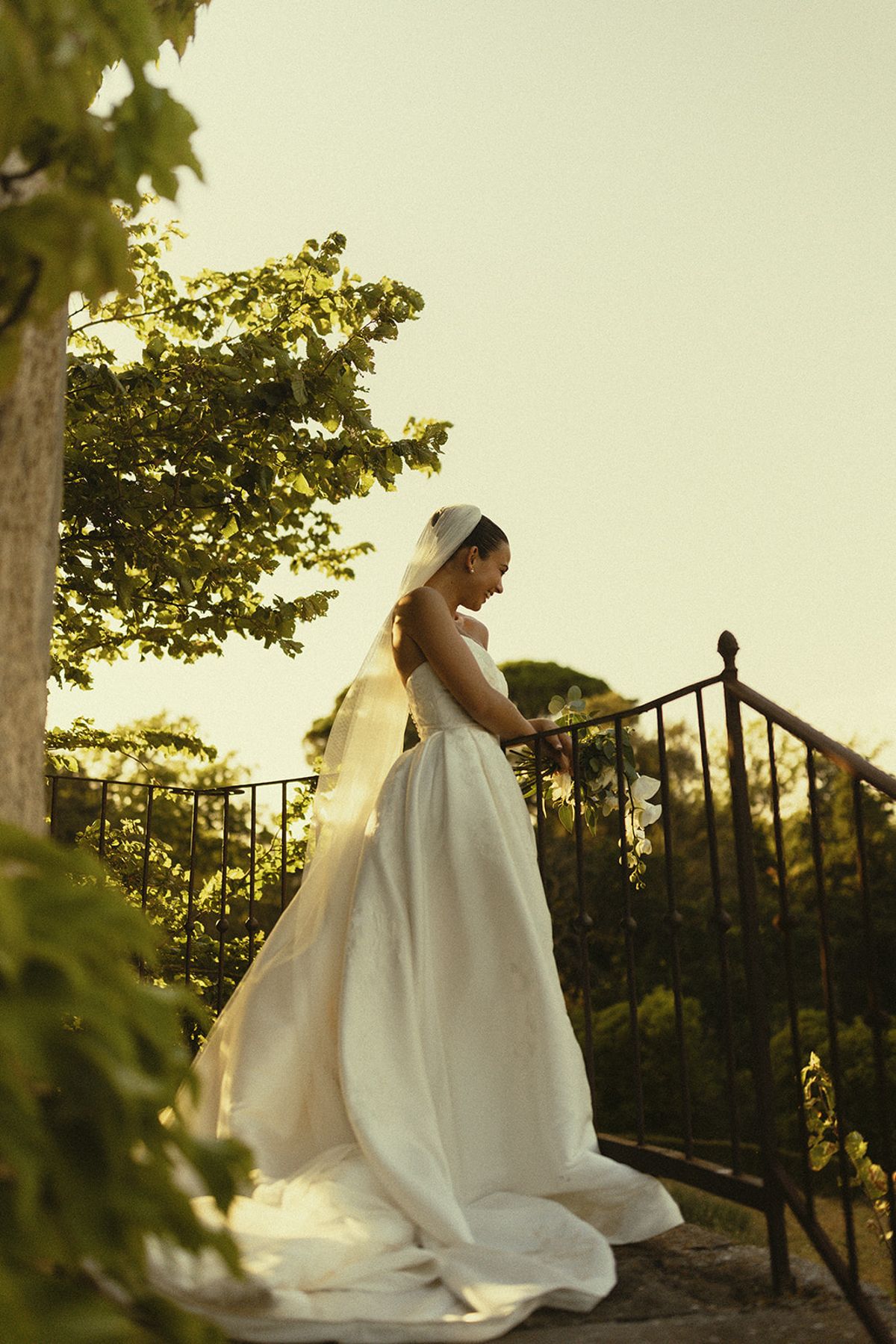The bride in a full white gown and long veil stands beside a black iron railing with trees behind her, lit by soft warm light.