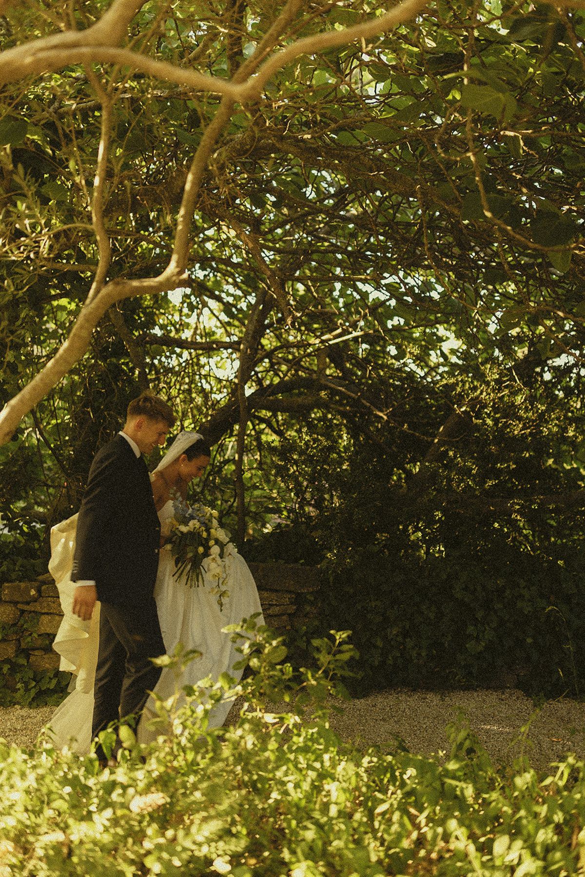 The groom in a dark suit stands beneath gnarled green trees, with the bride in white partly visible beside him.