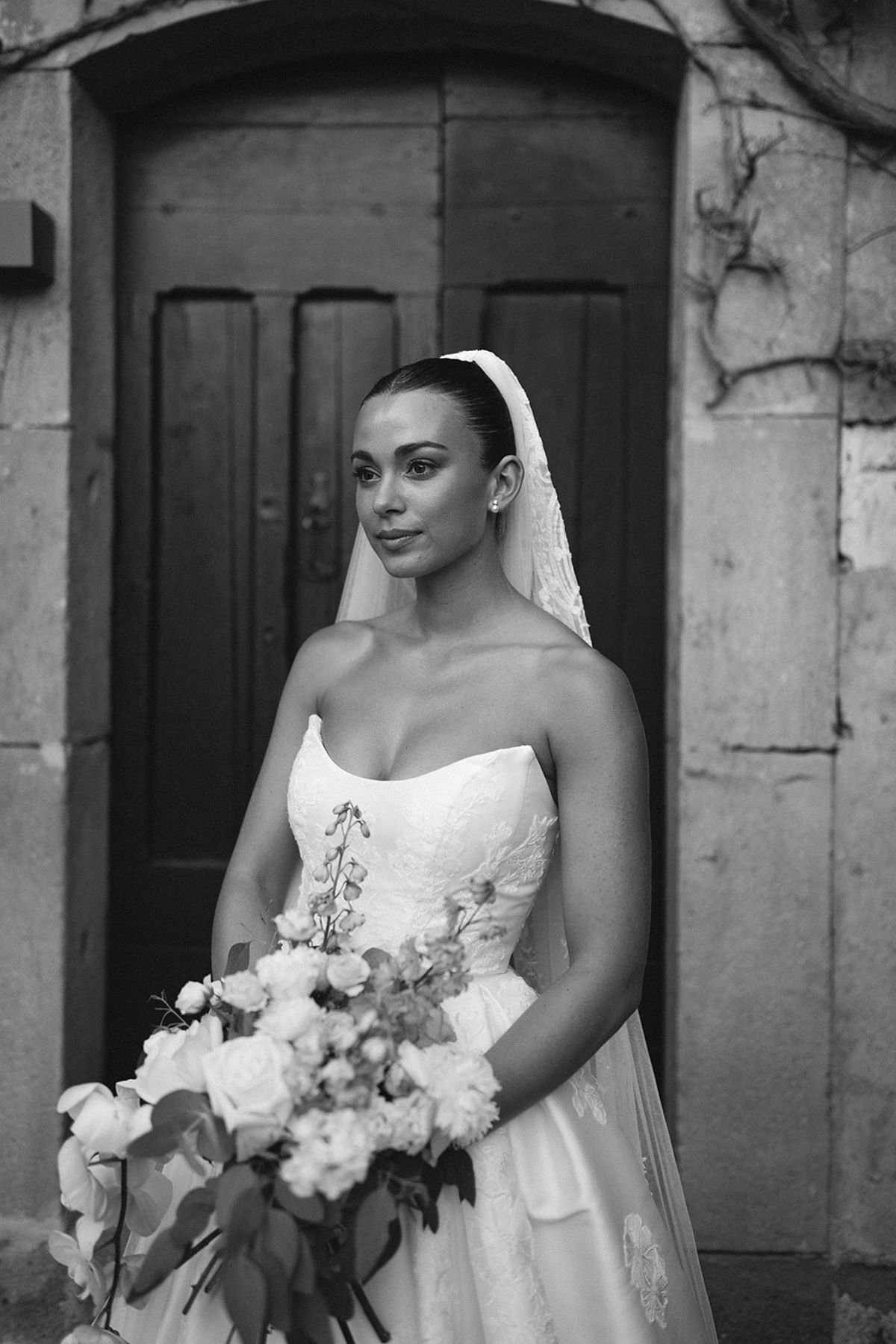In black and white, the bride in a veil and strapless gown holds a large bouquet in front of a wooden door.