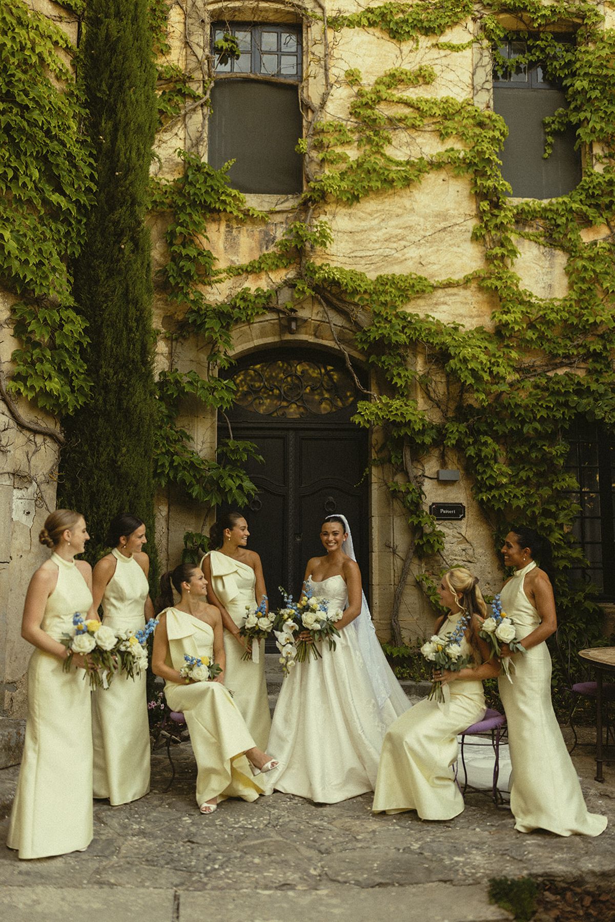 The bride and her bridesmaids in cream gowns gather in front of a stone building covered in ivy and tall cypress.