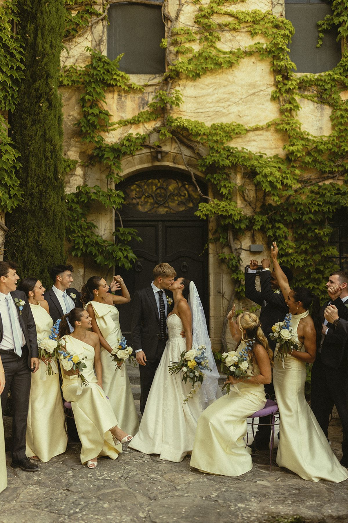 The couple stand with bridesmaids in pale dresses and groomsmen in suits, some cheering, in front of an ivy-covered doorway.