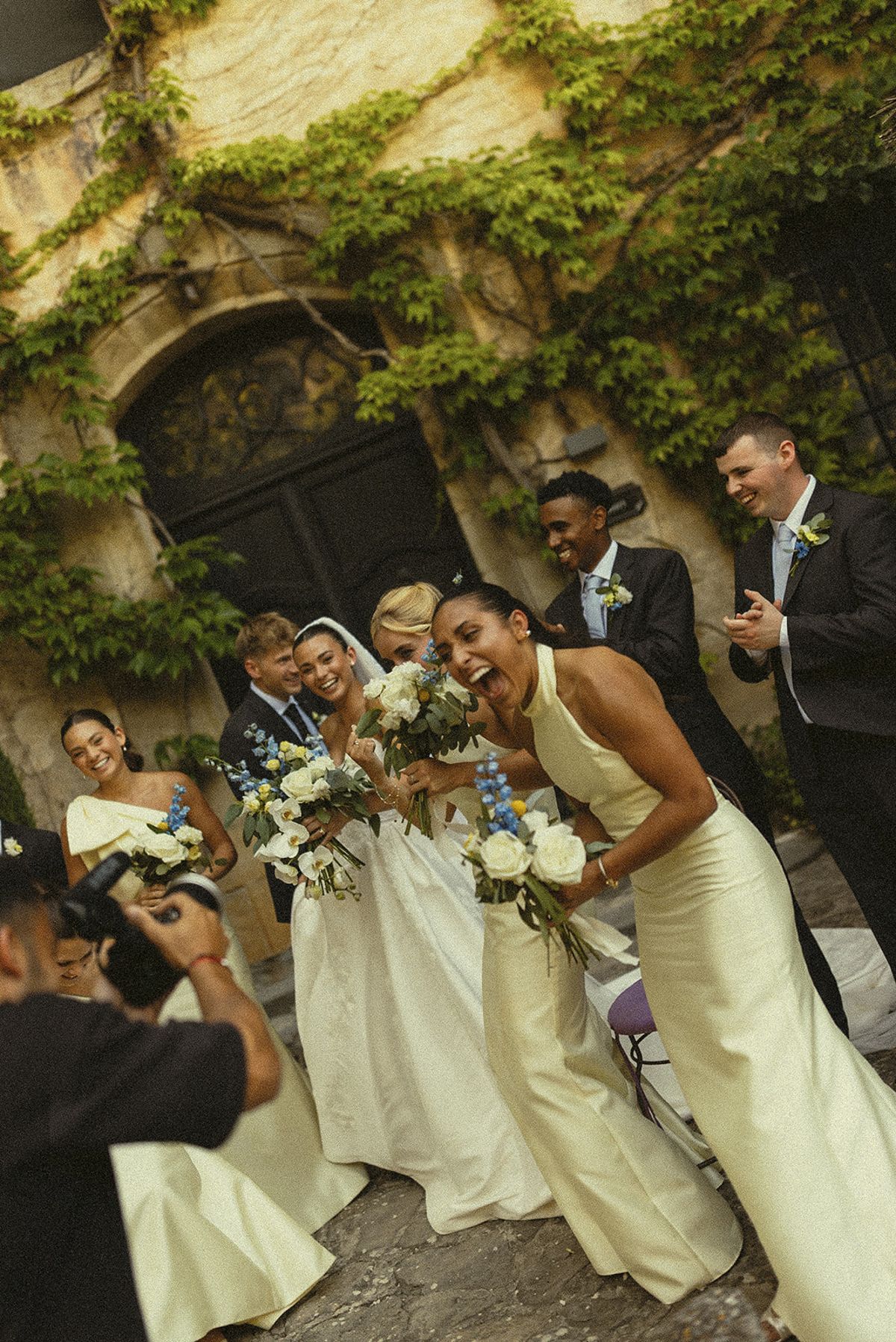 A group of women holding bouquets laugh together in a courtyard beneath an ivy-covered wall, with a photographer in the foreground.