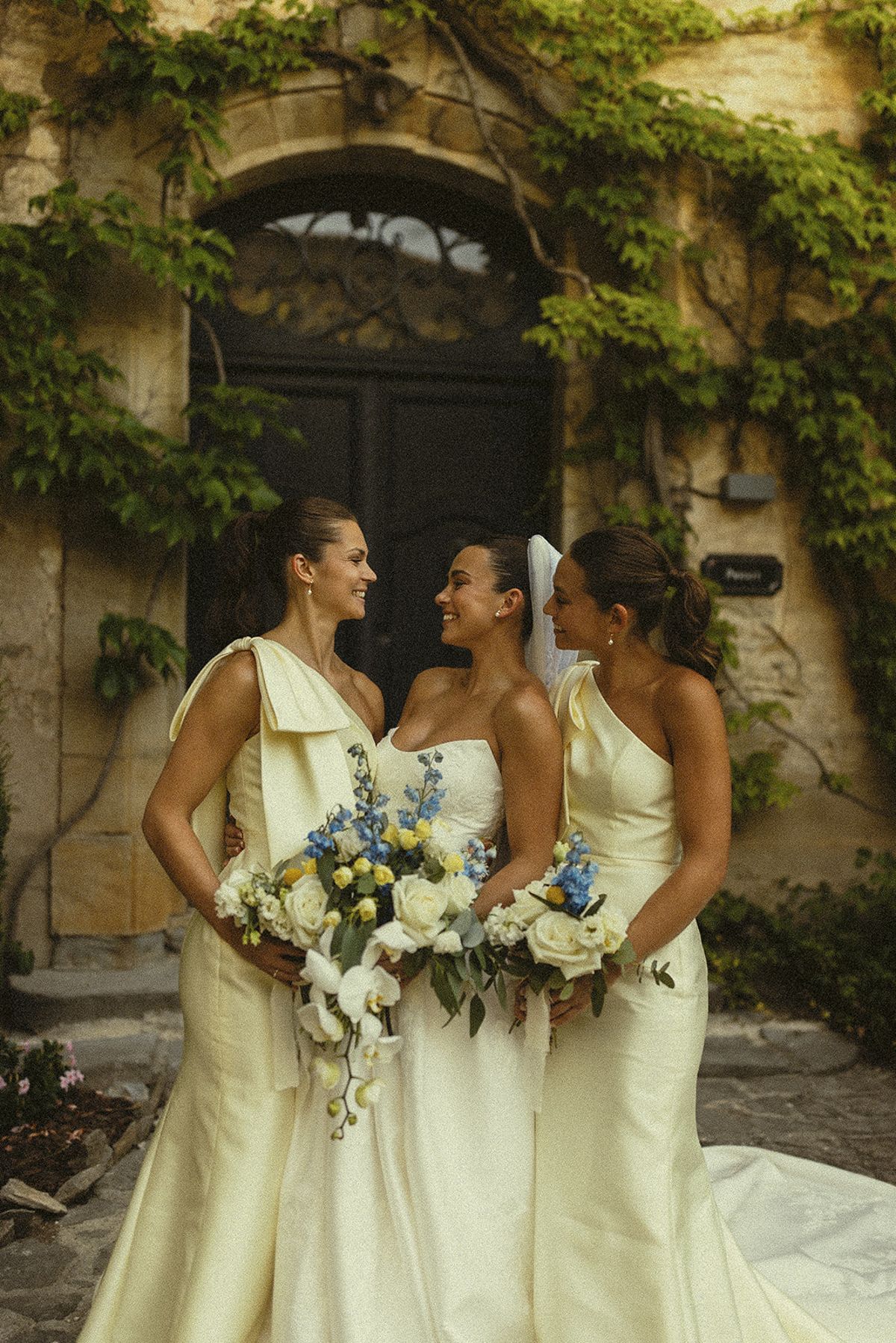 The bride stands between two bridesmaids in cream gowns, all holding bouquets, in front of an ivy-covered doorway.