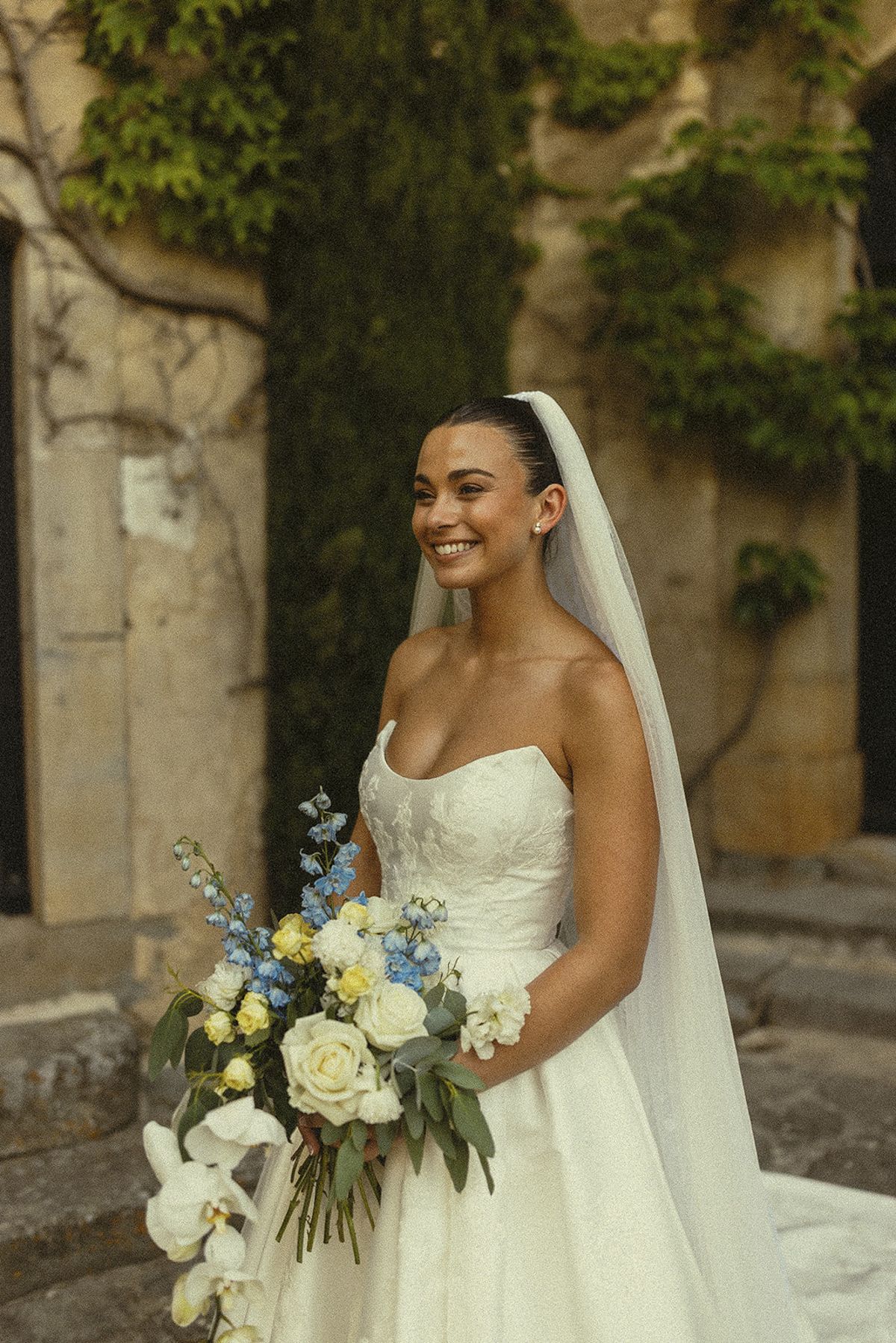 The bride smiles while holding a bouquet of white and blue flowers, standing before a stone wall covered in ivy.
