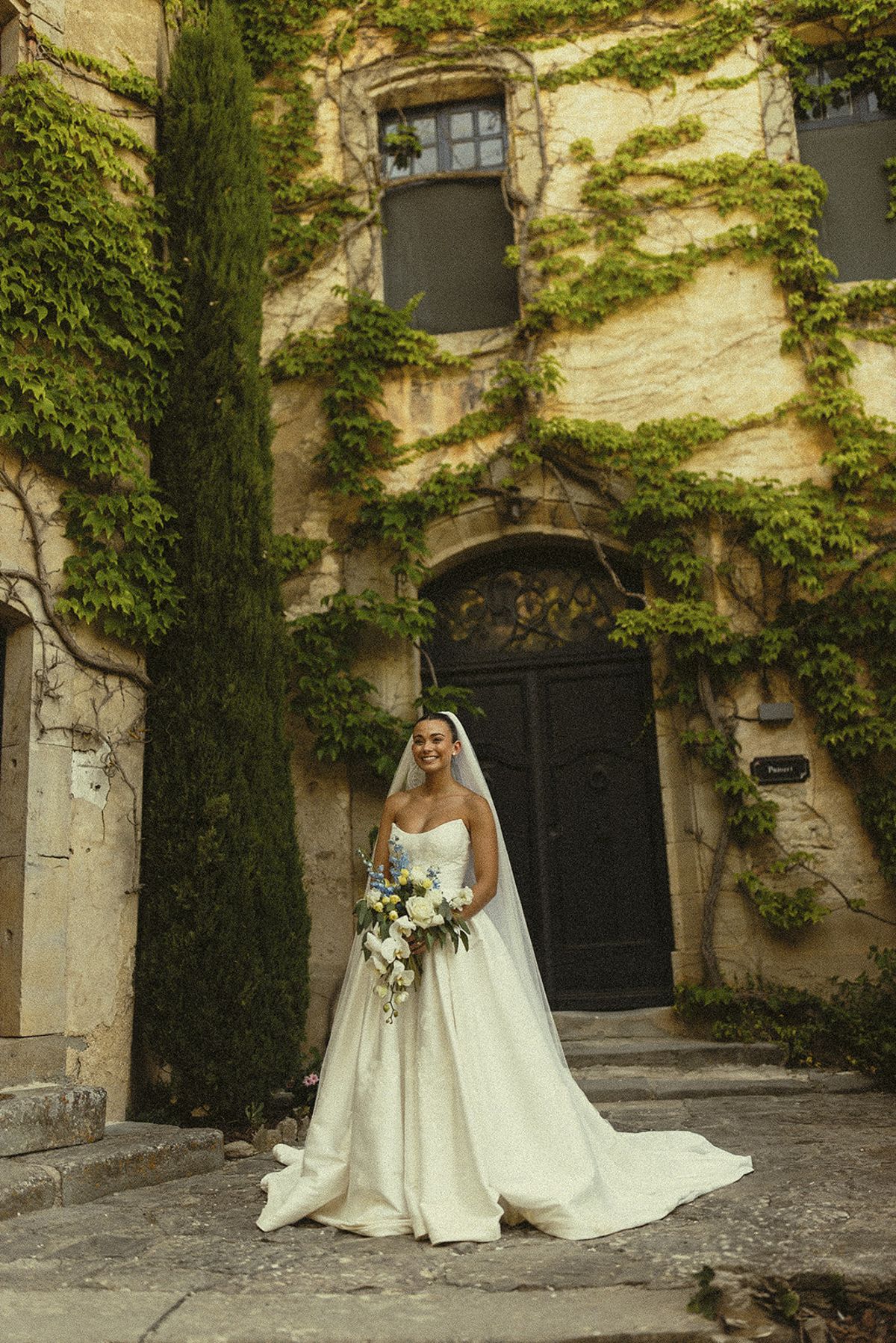 The bride in a full white gown holding a bouquet stands before a wooden door in an ivy-covered stone building beside a cypress.