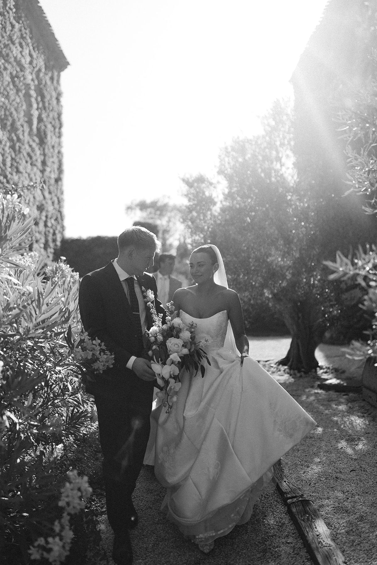 In black and white, the groom and the bride in a full gown walk along a garden path with bright sunlight behind.
