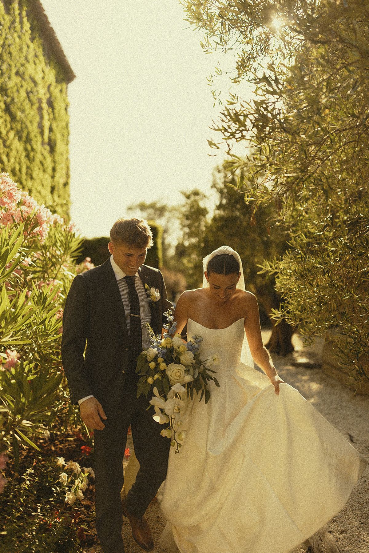The groom in a grey suit and the bride in a white gown walk side by side along a path lined with olive trees and flowers.