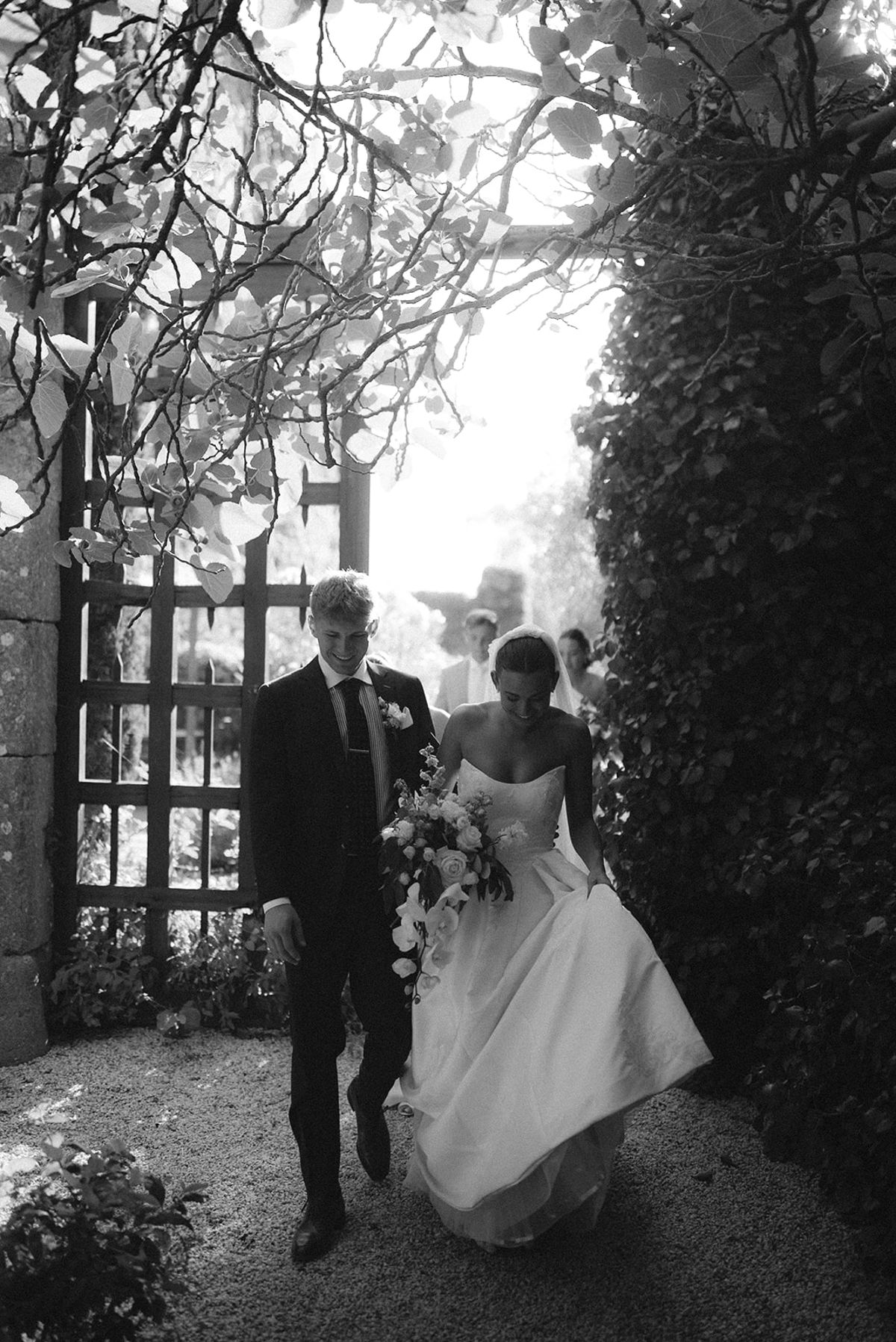 In black and white, the groom and the bride in her gown walk together through a wrought-iron gate in a leafy garden.