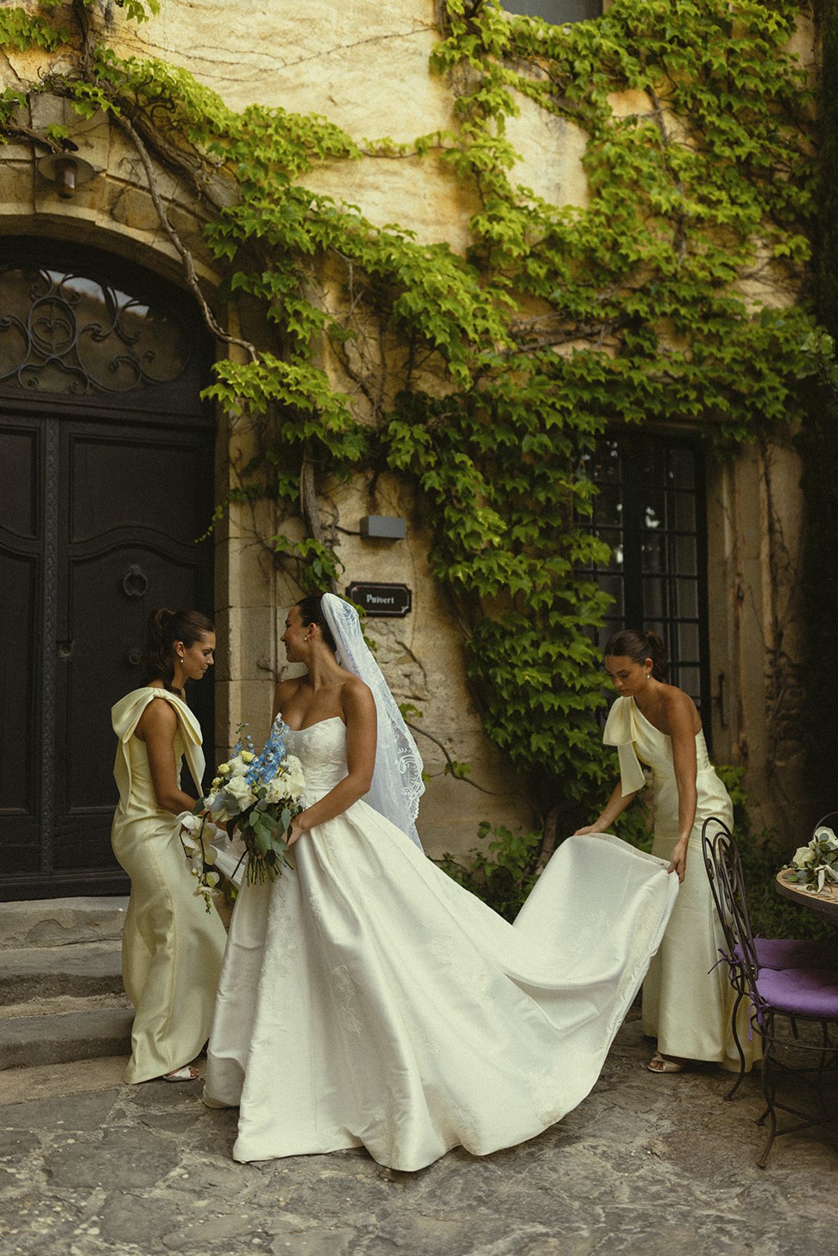 Two bridesmaids in pale yellow spread the long train of the bride's gown in front of an ivy-covered wall.