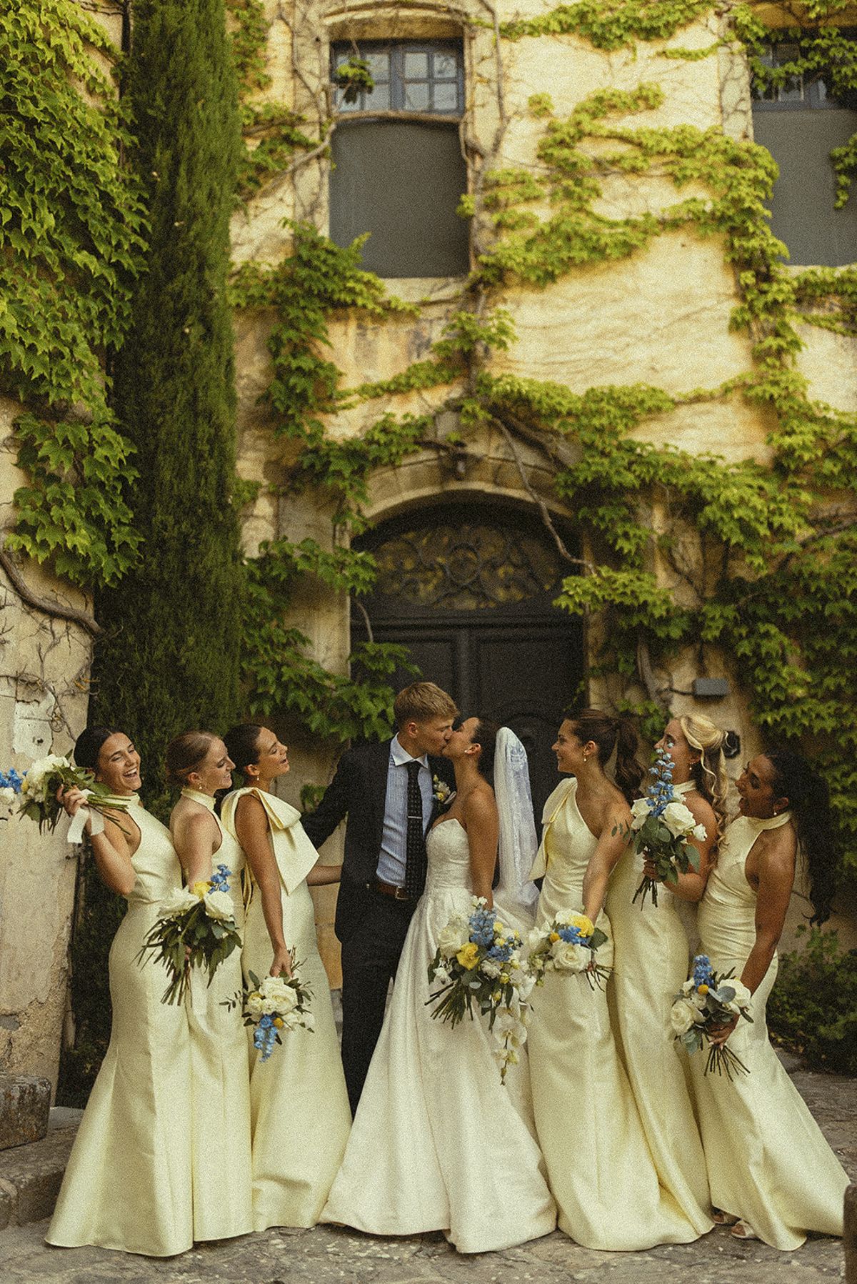 The couple stand among five bridesmaids in pale yellow dresses holding bouquets, in front of a stone building covered with green ivy.